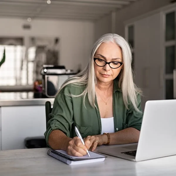 Mujer con gafas escribiendo en un cuaderno mientras está sentada a una mesa con un portátil.