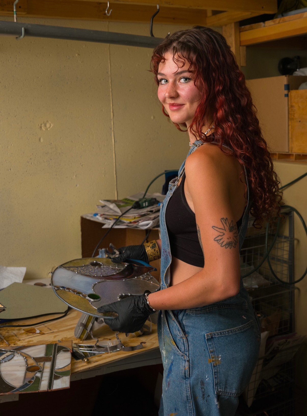 Young woman with curly red hair and a tattoo on her arm, wearing a black sleeveless top and denim overalls, standing in a workshop holding a stained glass art piece.