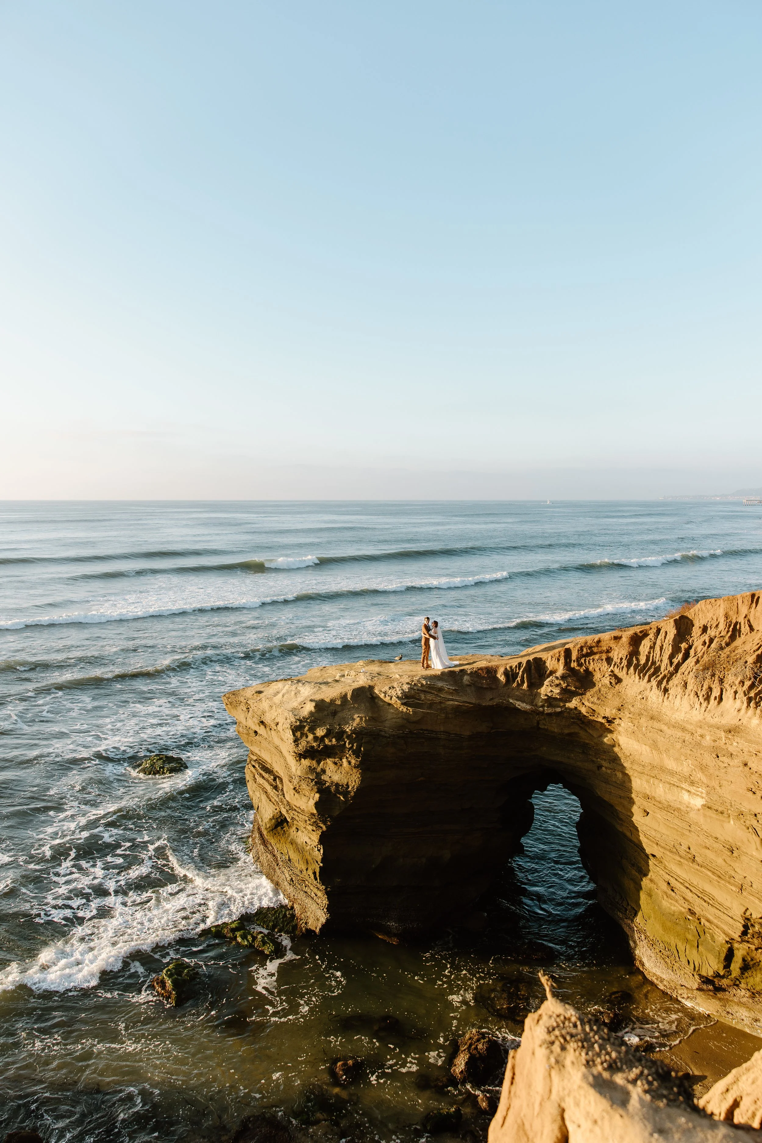 A couple dressed in wedding attire stands on a rock formation near the ocean, with a large natural arch below them and the sea in the background during sunset.