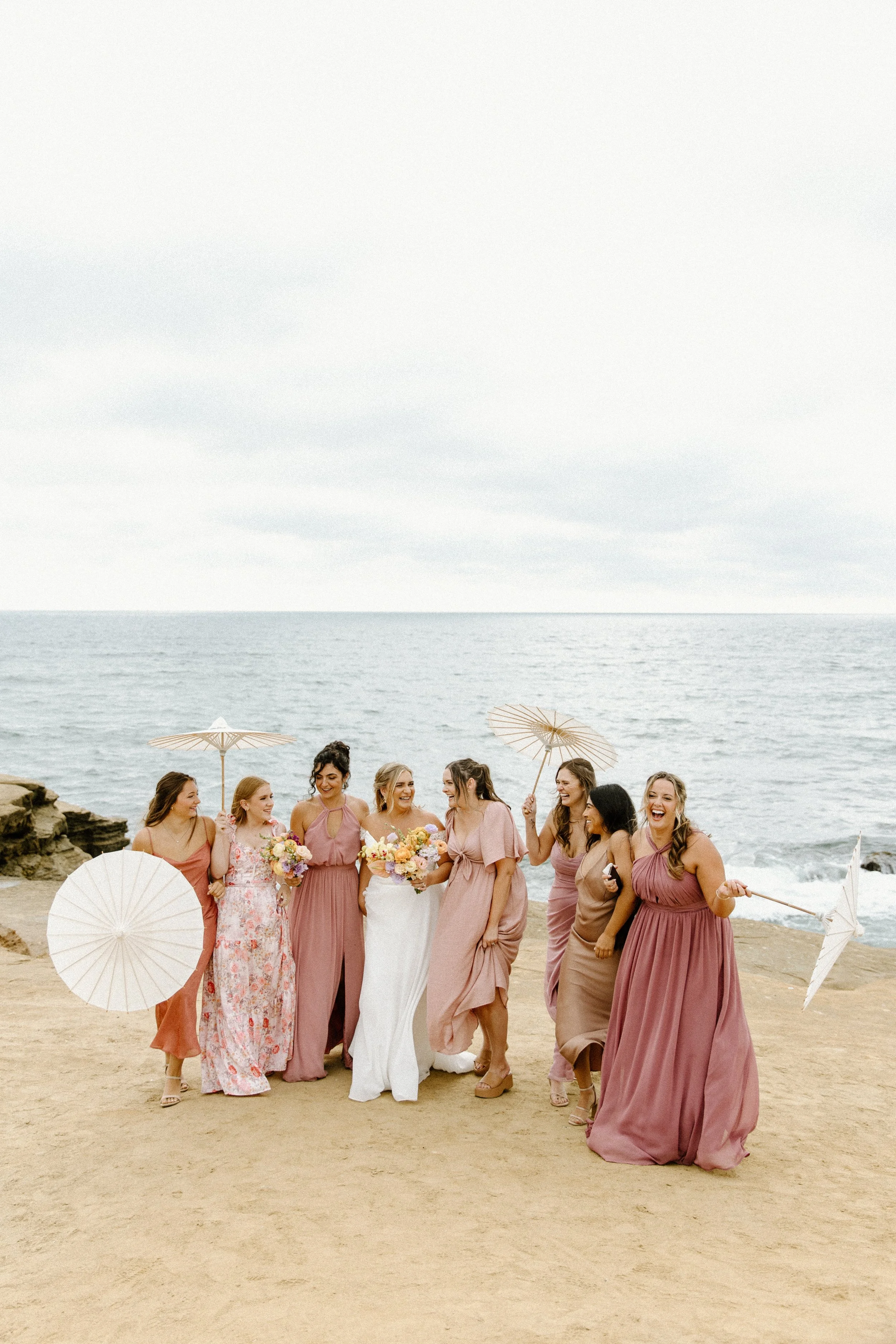 Group of women celebrating at a beach, dressed in pink and neutral dresses, holding parasols and bouquets, with the ocean in the background.