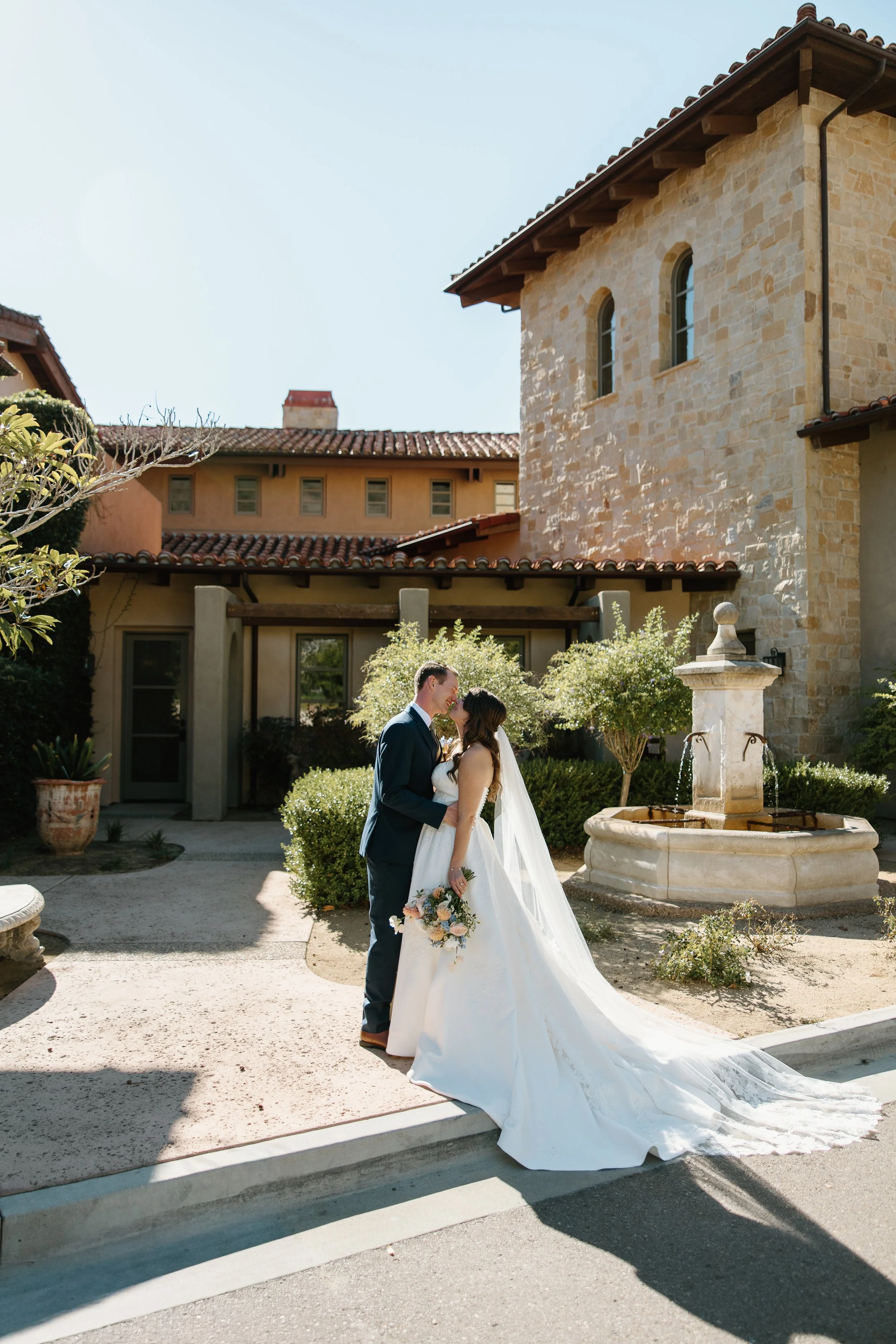 A bride and groom share a kiss outdoors on a sunny day at a wedding, with the bride in a white gown and veil holding a bouquet, and the groom in a dark suit, in front of a stone building and a fountain.