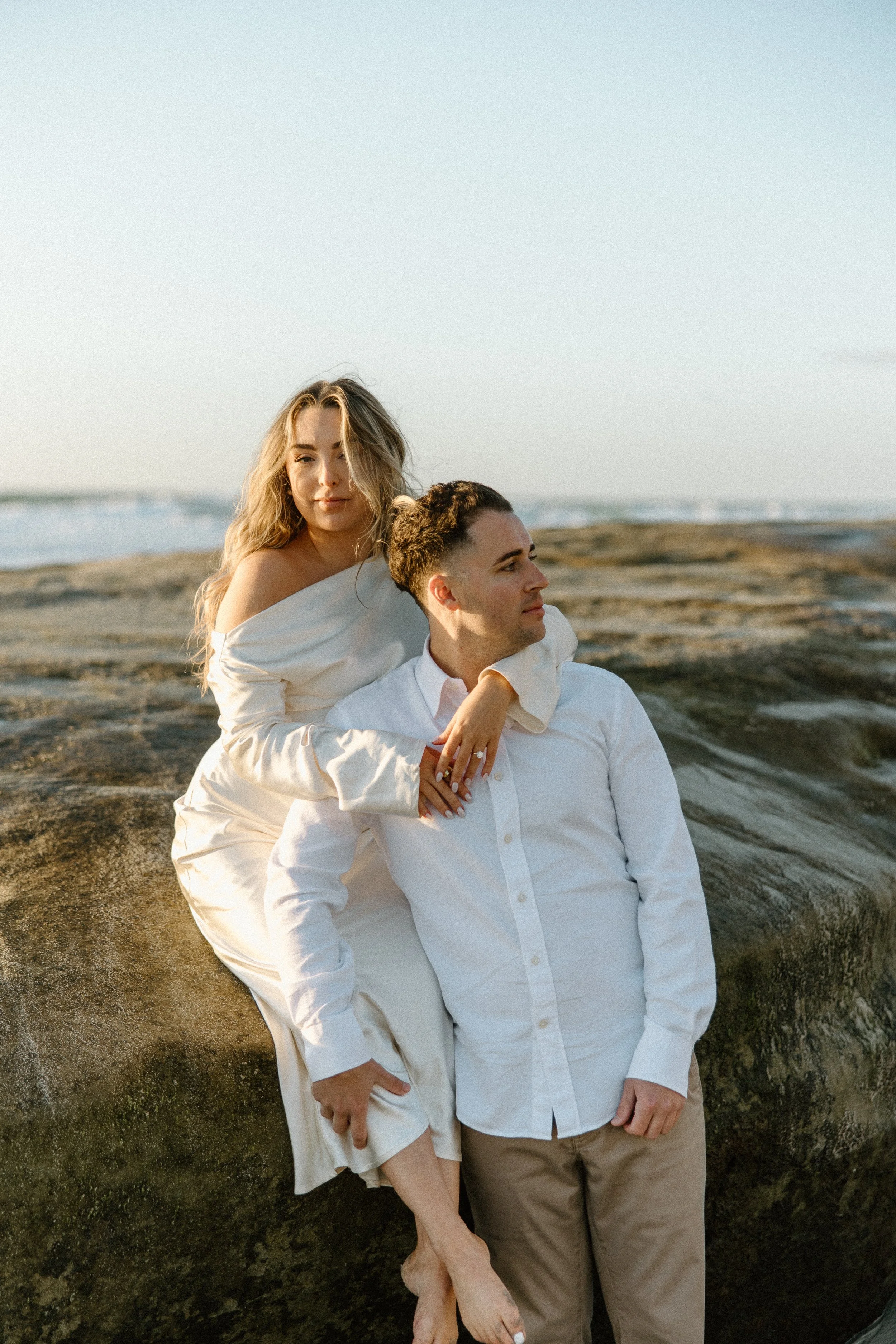 A woman and a man pose on a rocky beach at sunset, with the woman sitting on a large rock and the man standing beside her. Both are dressed in white and beige, looking contemplative against a backdrop of the ocean and sky.