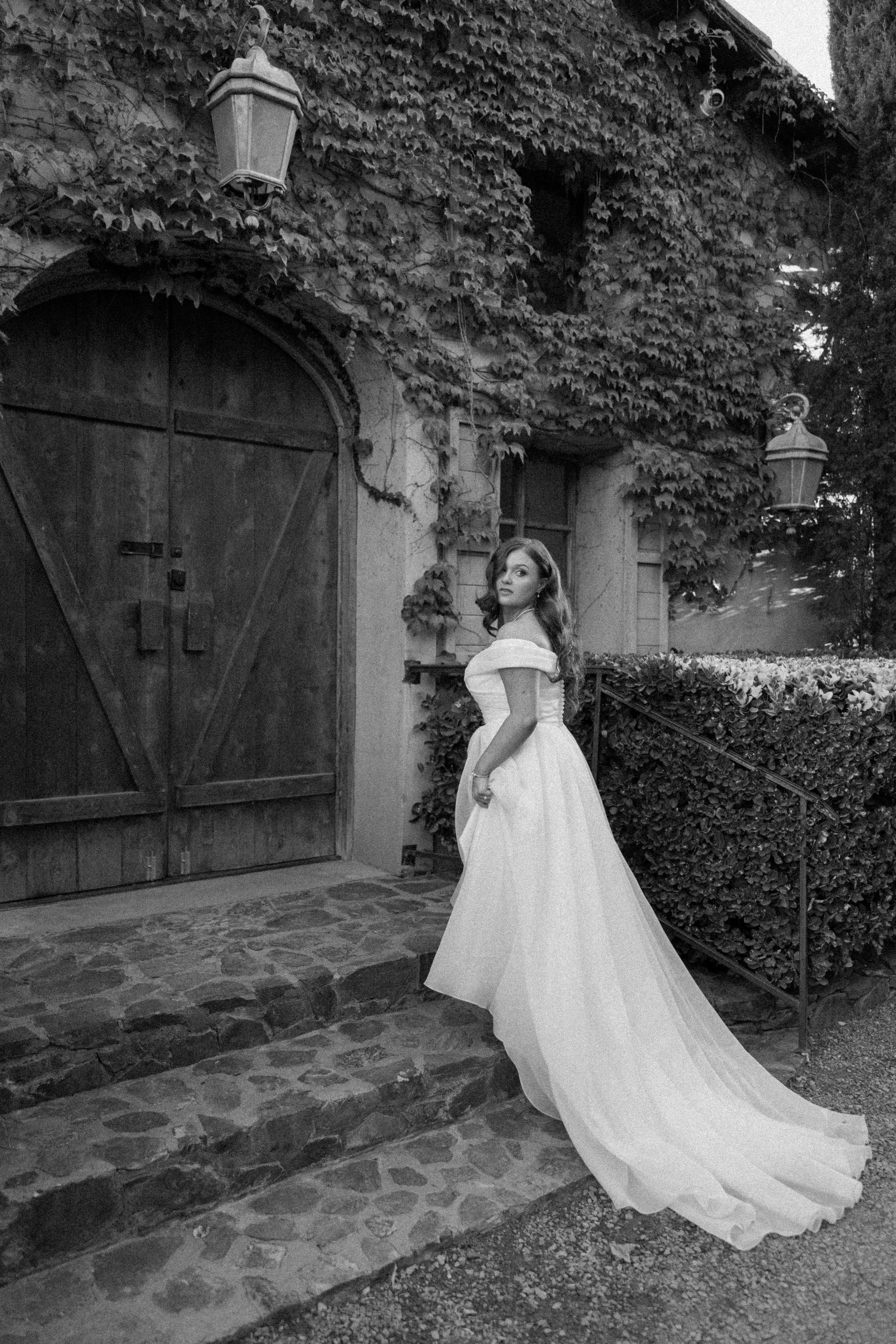 A woman in an elegant white gown stands on stone steps outside a rustic building covered in ivy, with wooden doors and lanterns.