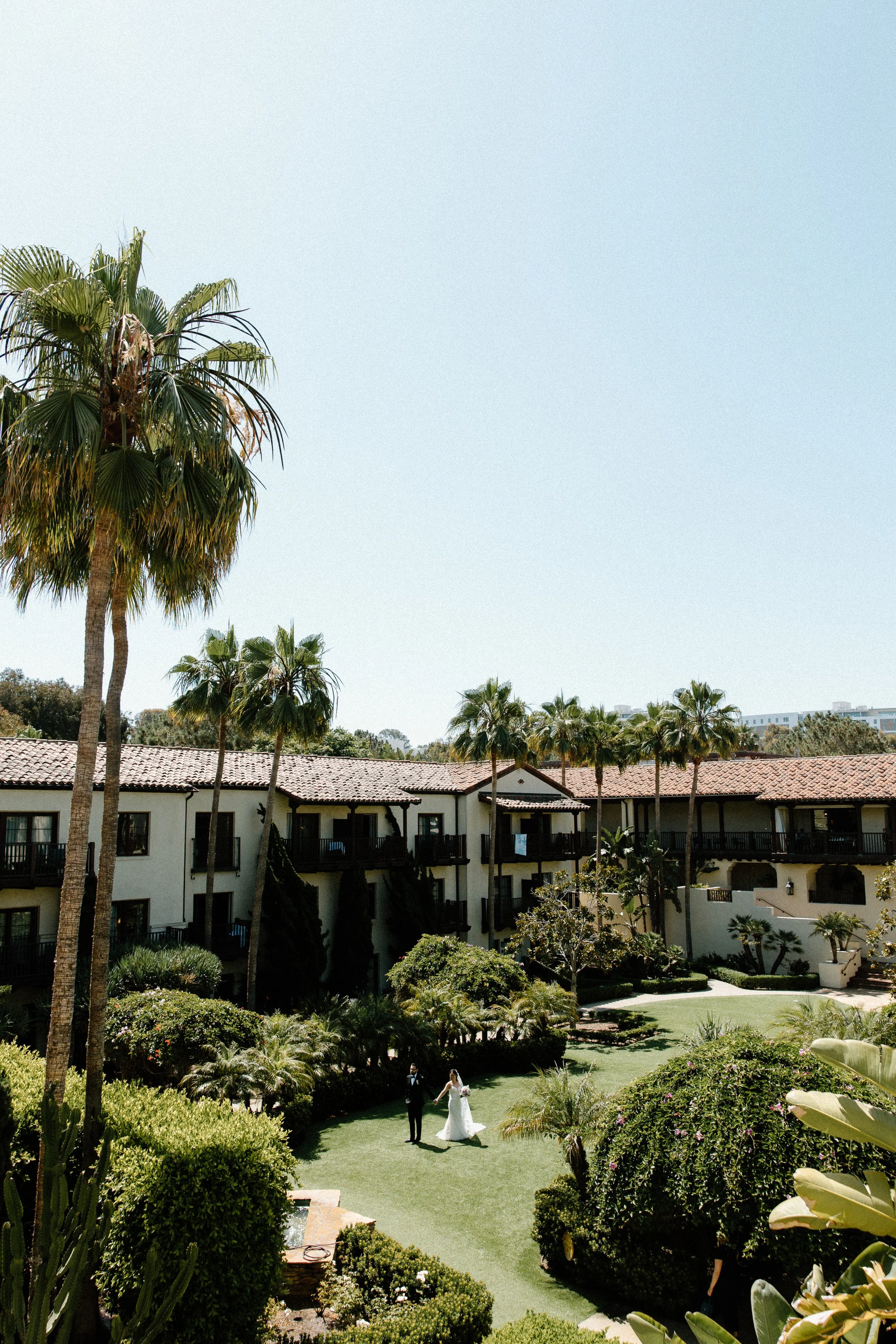 A couple in wedding attire walking in a lush, green garden courtyard with palm trees, surrounded by white residential buildings with red tile roofs, under a clear blue sky.