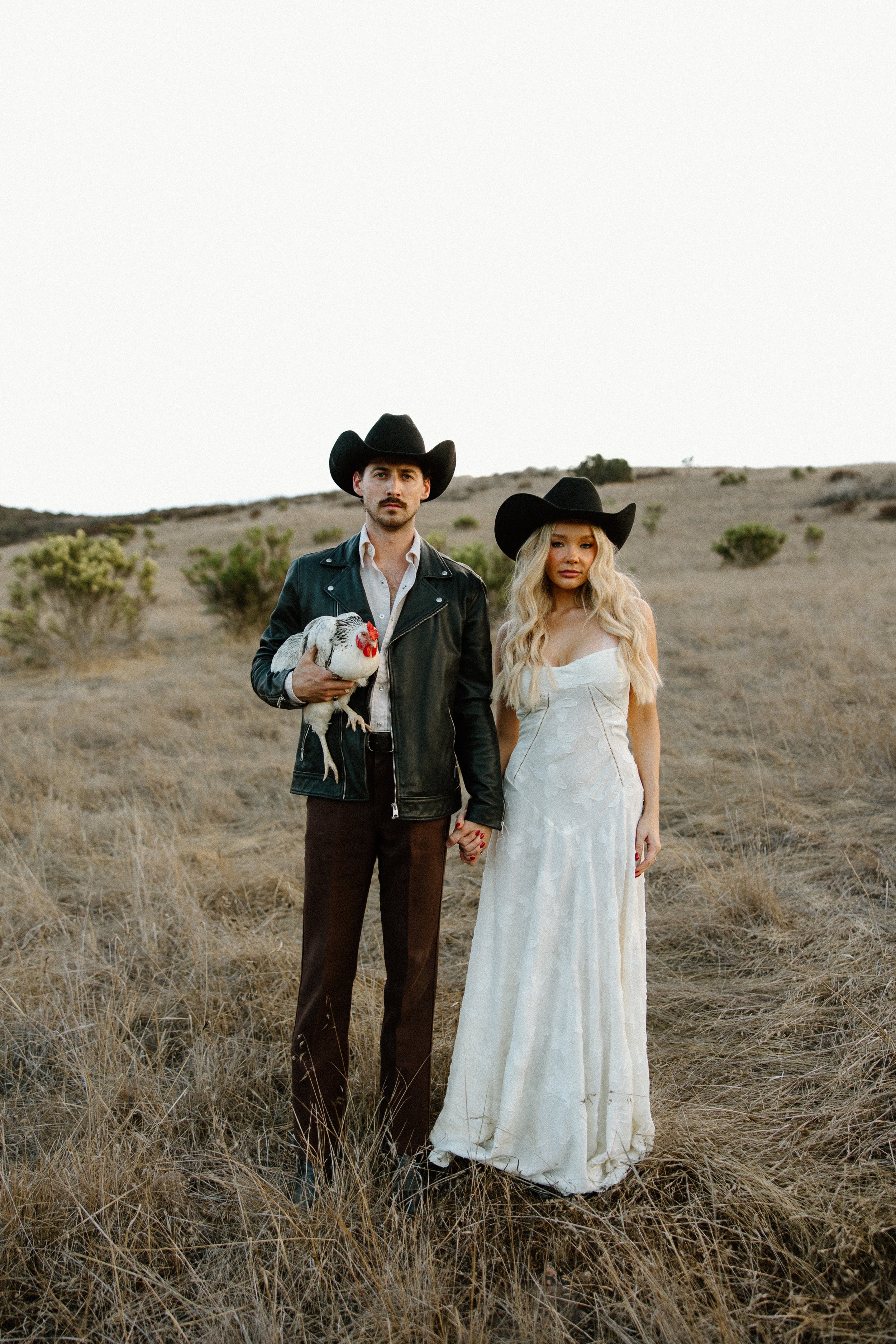A couple dressed in Western attire holding hands in a dry, grassy field with shrubs, the man holding a chicken, both wearing black cowboy hats, with a mostly clear sky in the background.