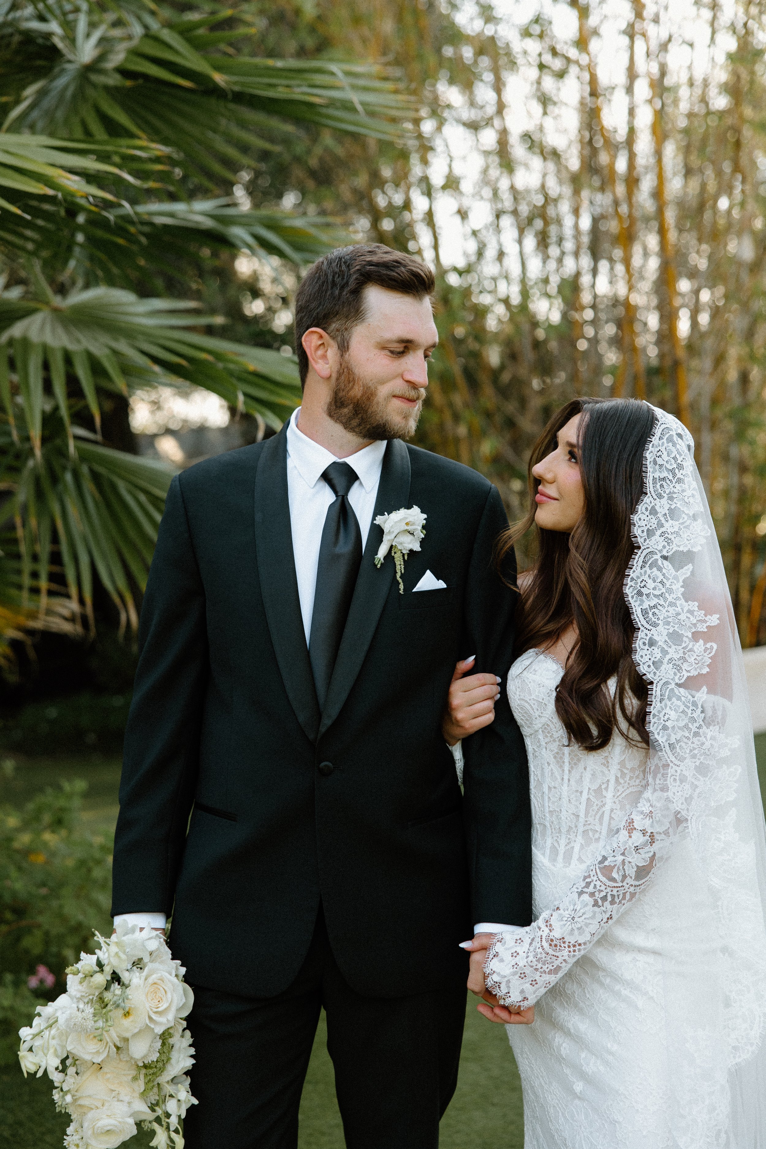 A bride and groom holding hands outdoors during their wedding, with the bride in a lace wedding gown and veil, and the groom in a tuxedo with a white flower boutonniere.