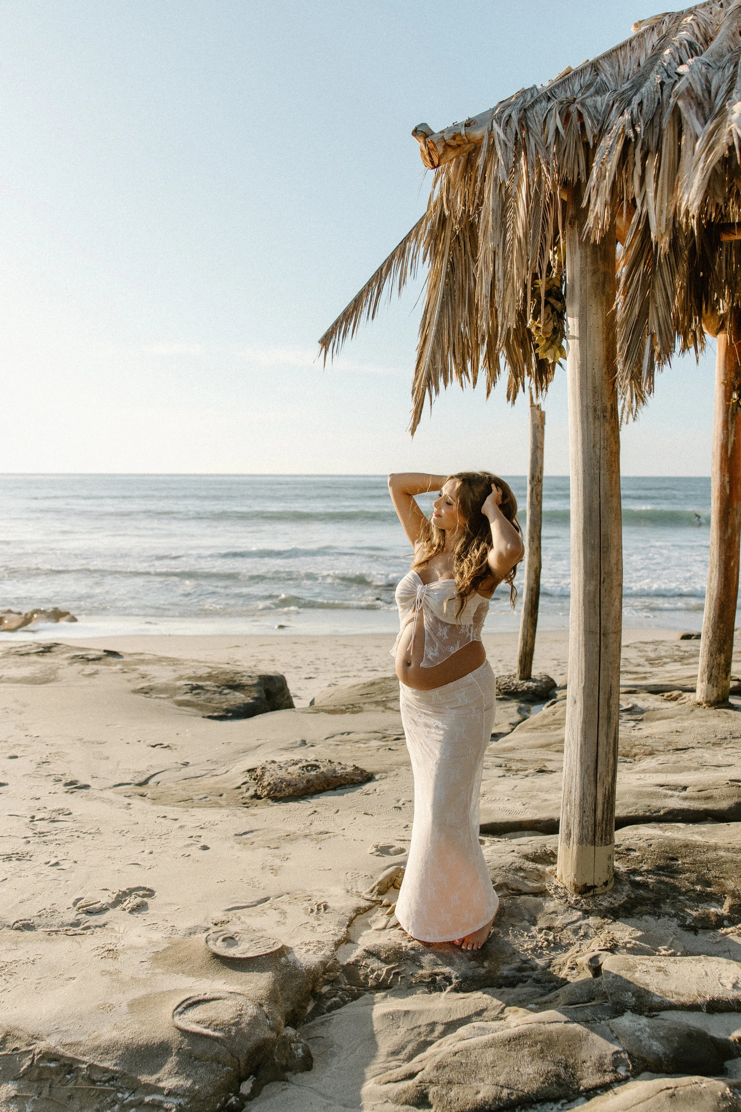 A pregnant woman in a white lace dress standing on a sandy beach under a thatched-roof hut, with the ocean in the background.