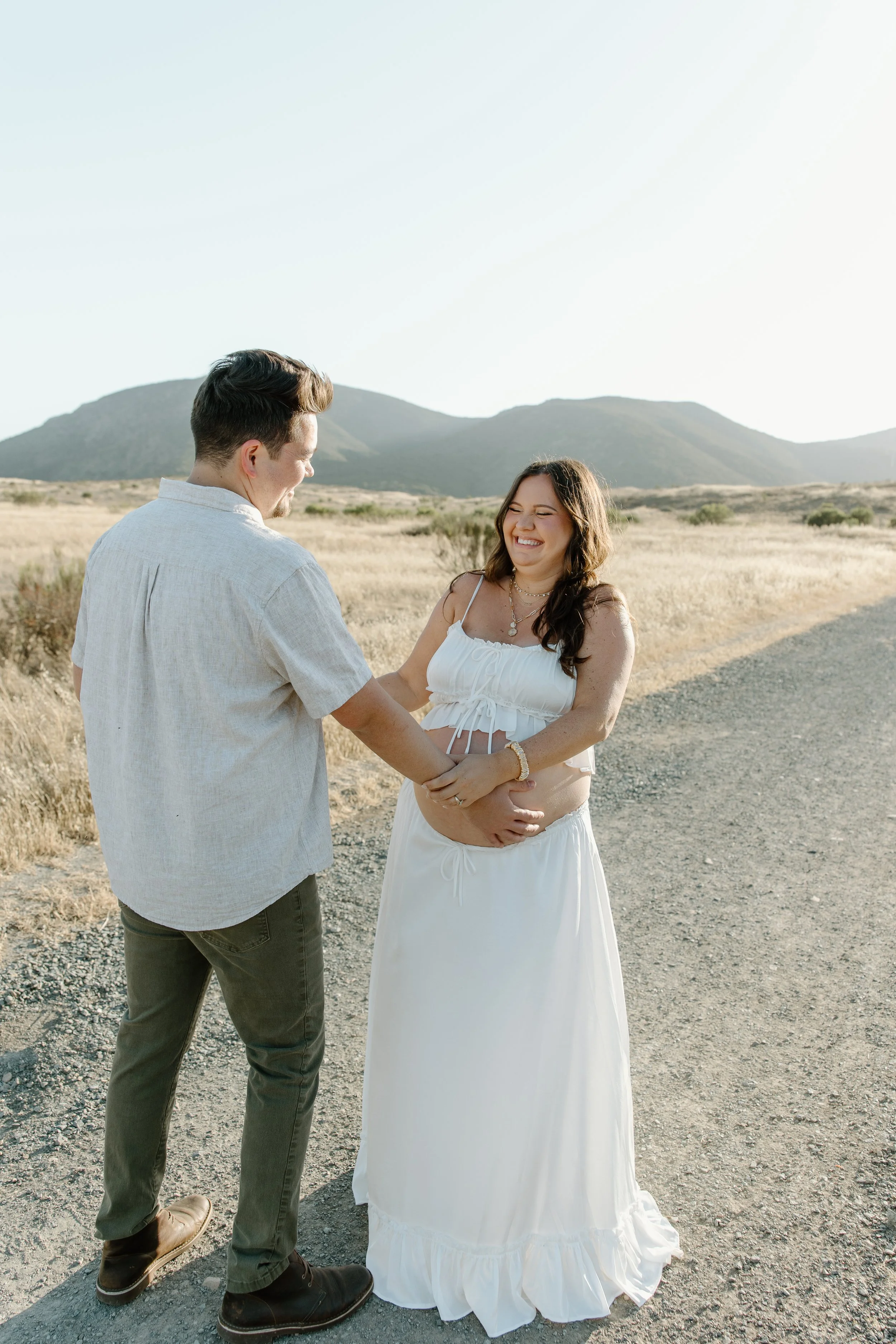 A man and a woman are standing on a dirt road in a desert landscape with mountains in the background. The man is holding the woman's hands, and they are both smiling and laughing. The woman is wearing a white, sleeveless dress, and the man is dressed