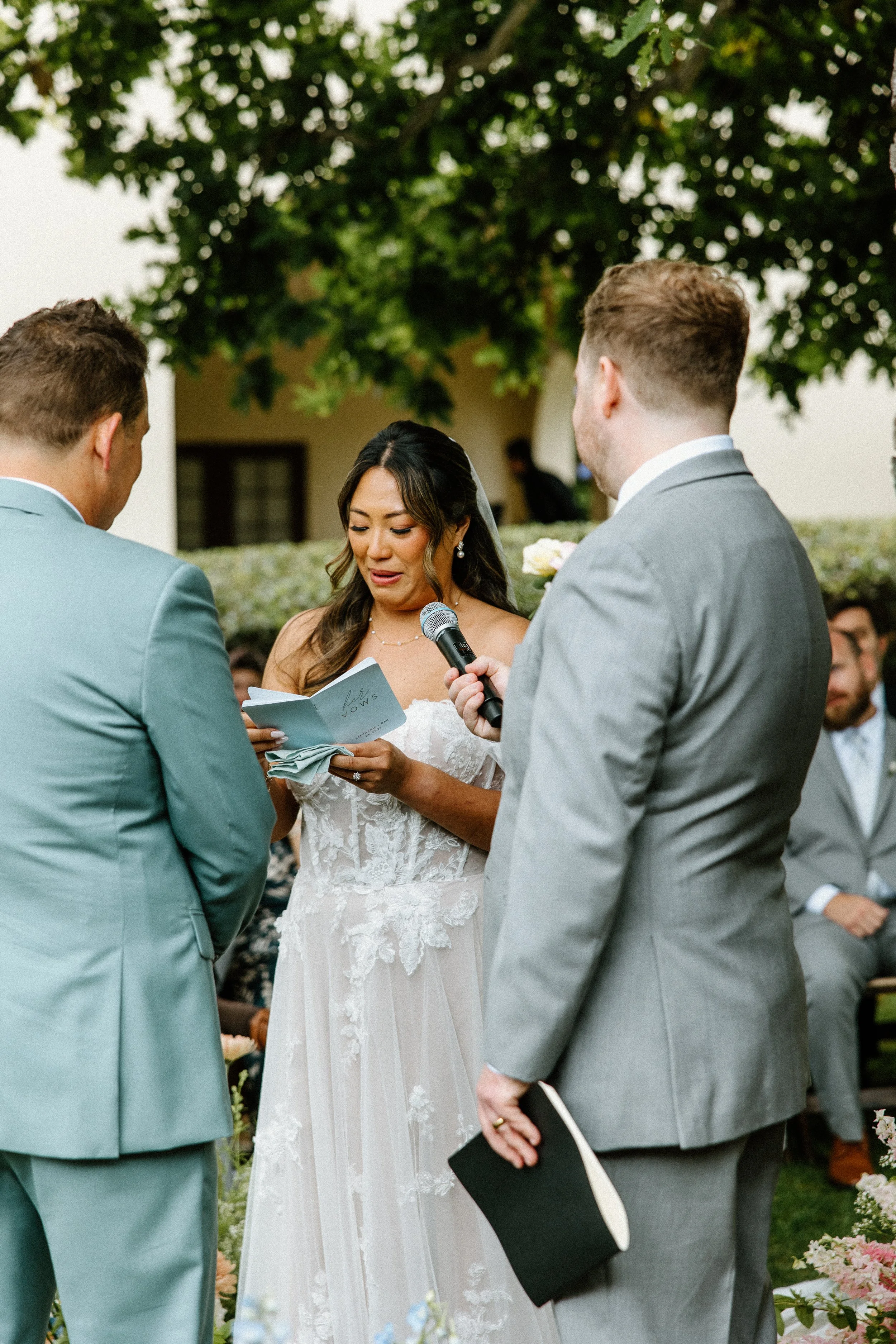 A bride and groom exchanging vows in an outdoor wedding ceremony, with officiant reading vows from a booklet and holding a microphone, in front of seated guests and trees.