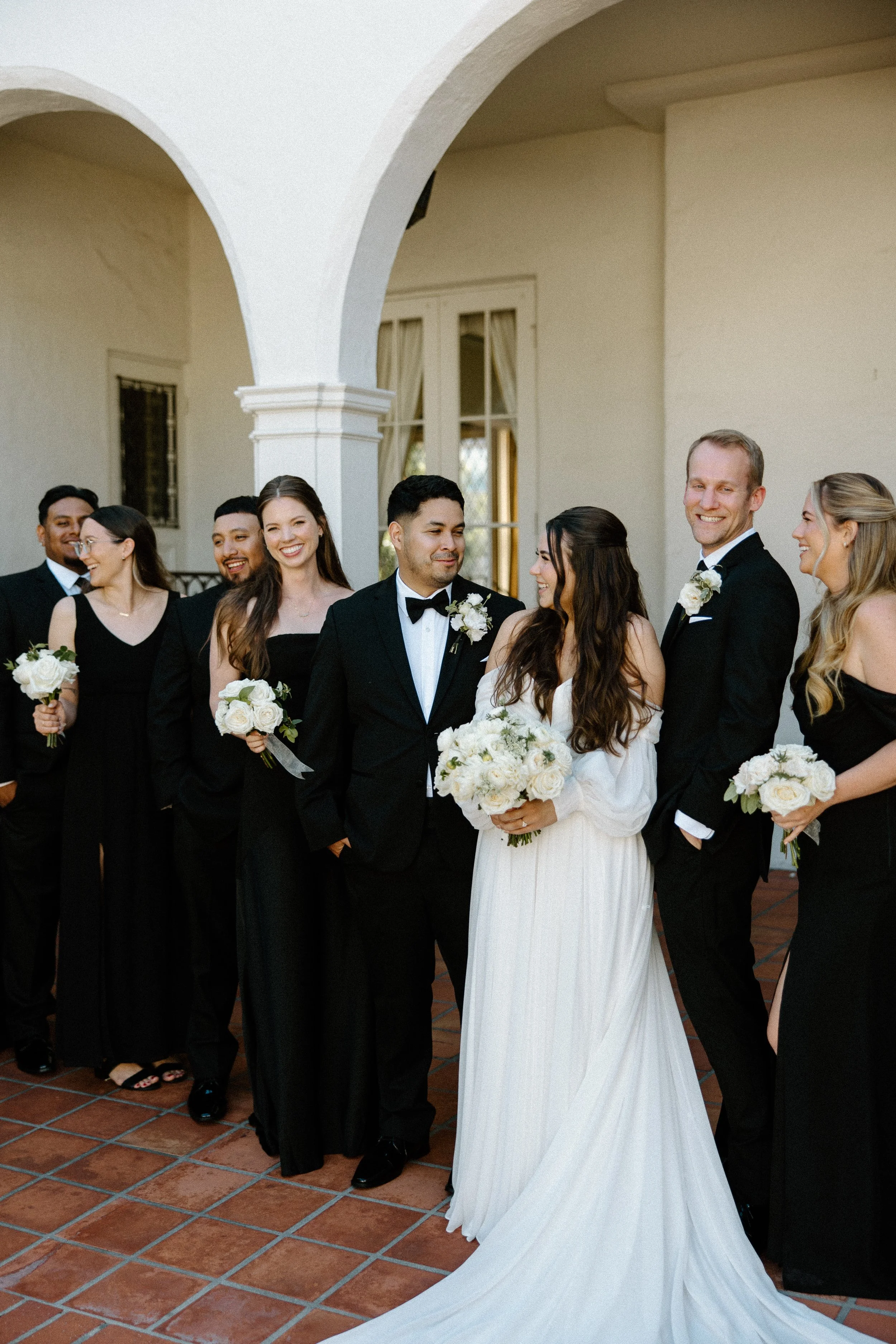 Group of wedding guests standing together under an archway, dressed in formal attire, with a bride holding a bouquet in the center.