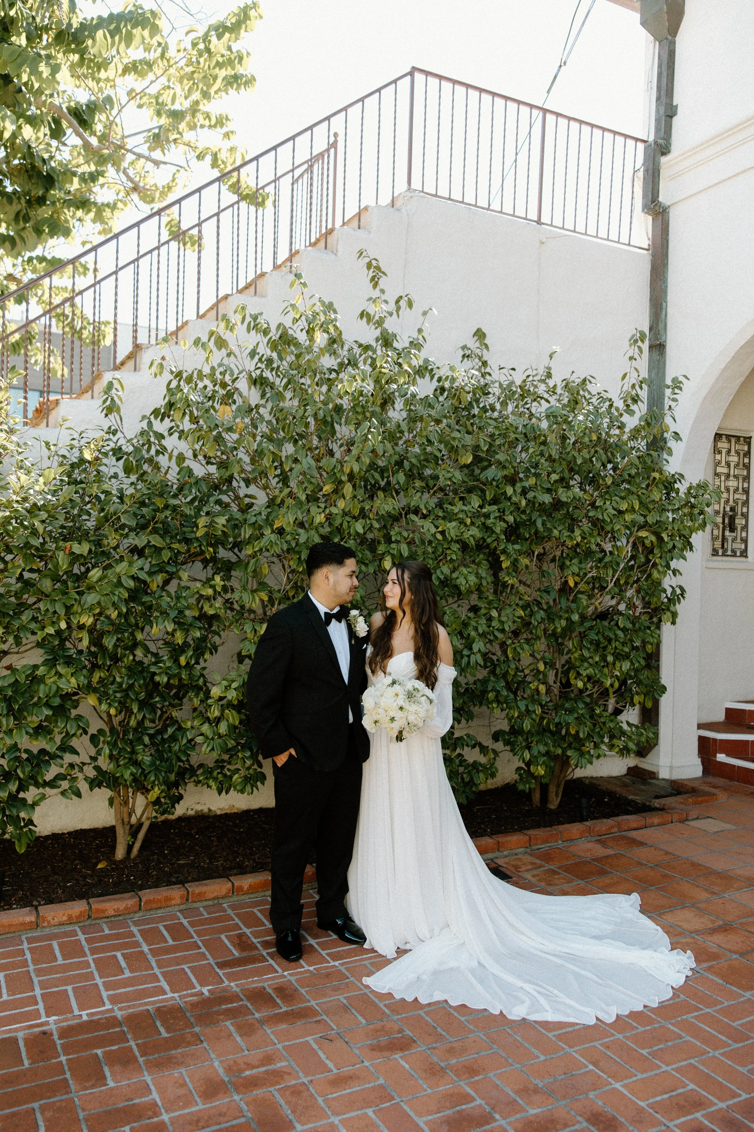 A bride and groom stand on a brick patio, gazing into each other's eyes, with a leafy green bush behind them and a white building with stairs and a railing in the background.