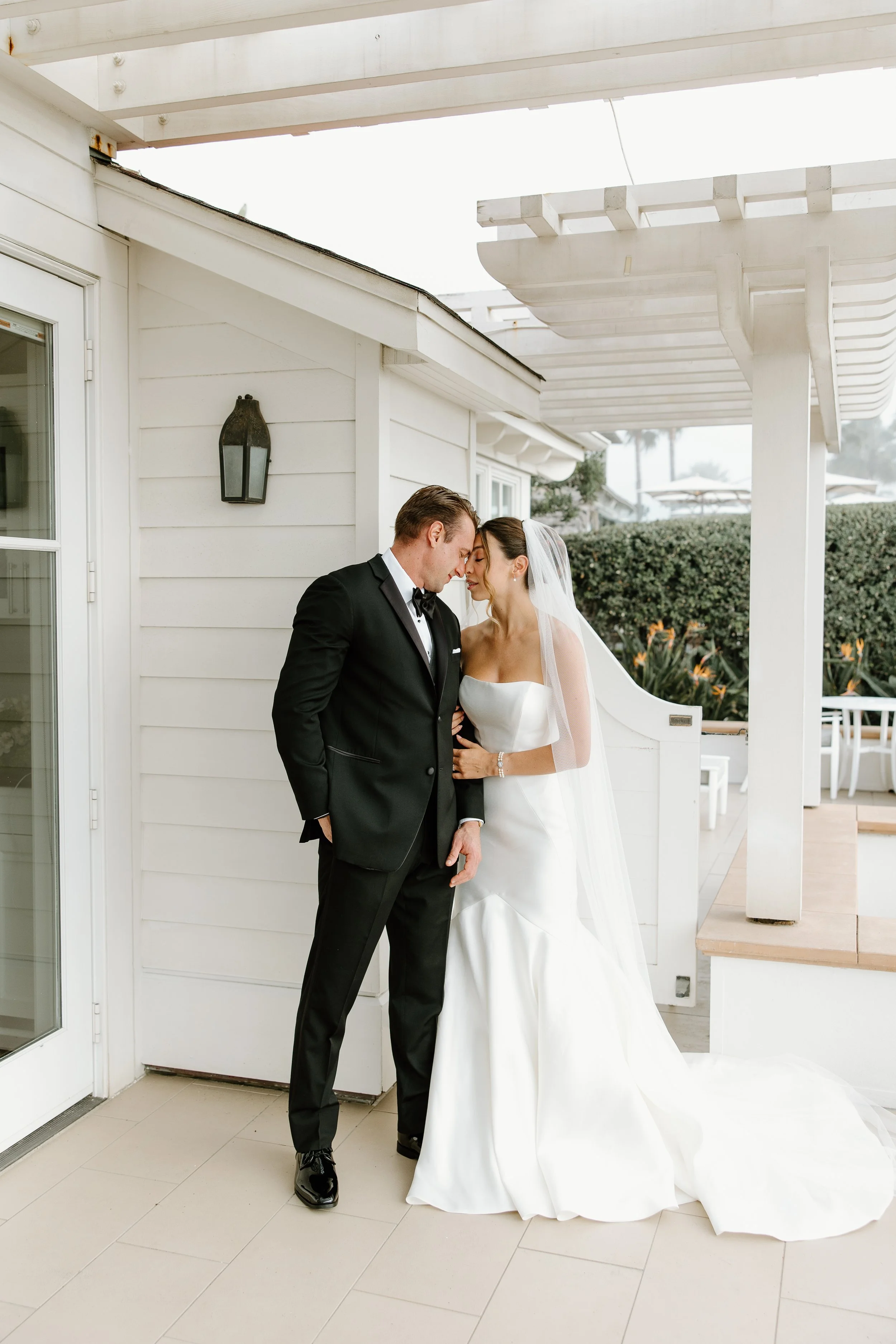 A bride and groom standing close together outside a white house, with the bride wearing a strapless white wedding dress and veil, and the groom in a black tuxedo, touching foreheads in an intimate moment.