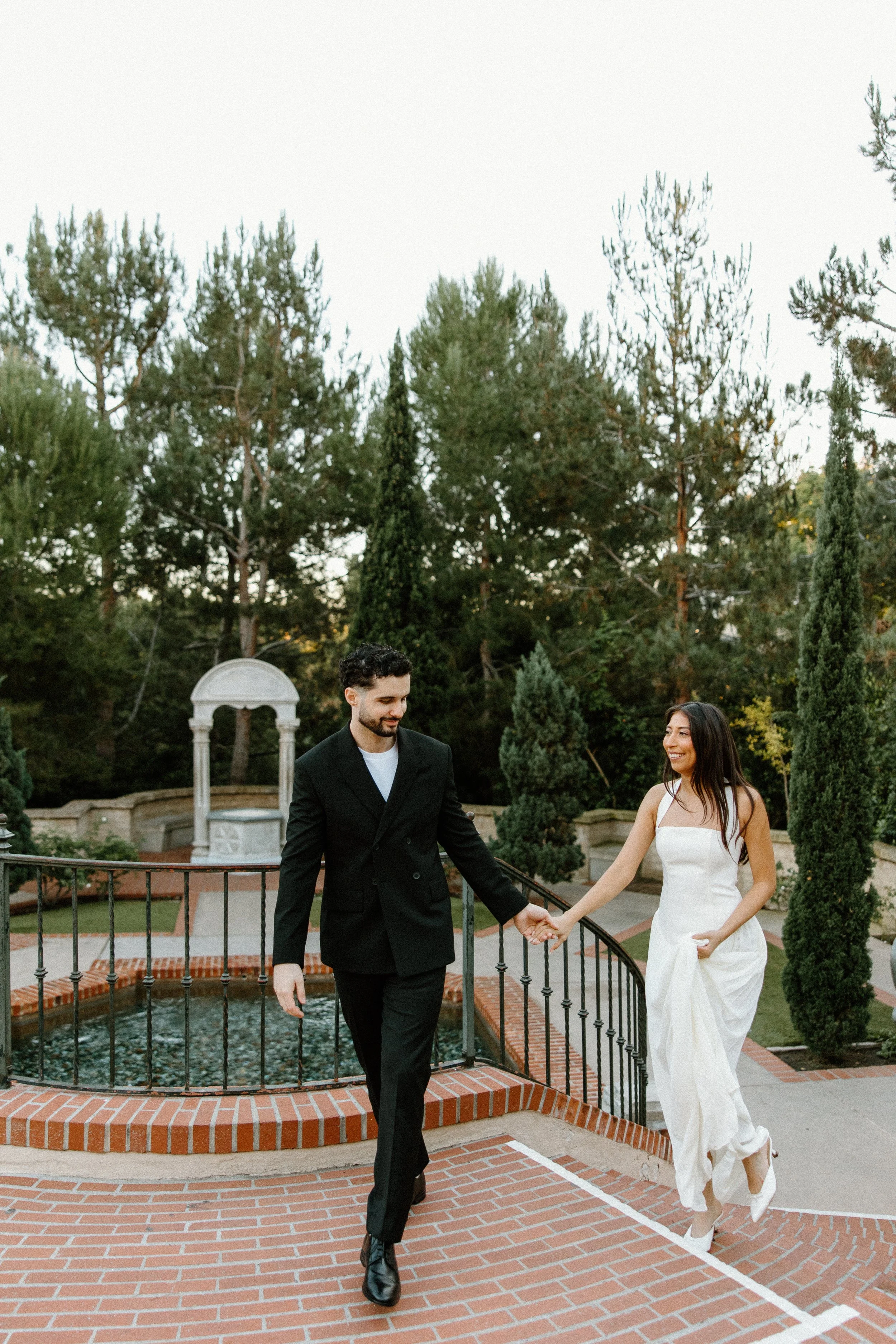 A couple holding hands and walking down a brick pathway in a garden, with the woman in a white dress and the man in a black suit, surrounded by trees and greenery.