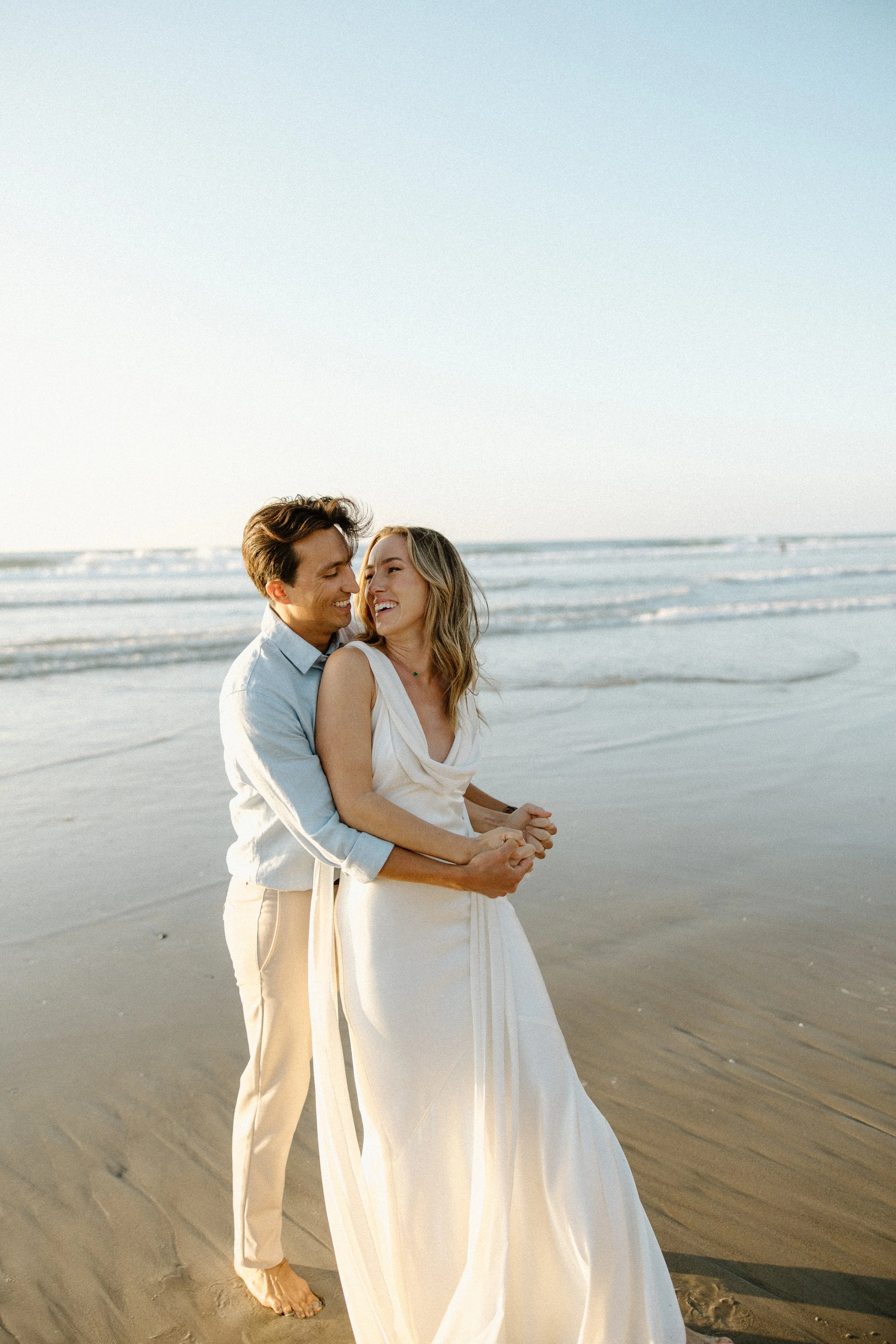 A joyful couple standing on a sandy beach with the ocean waves in the background, embracing each other and smiling.