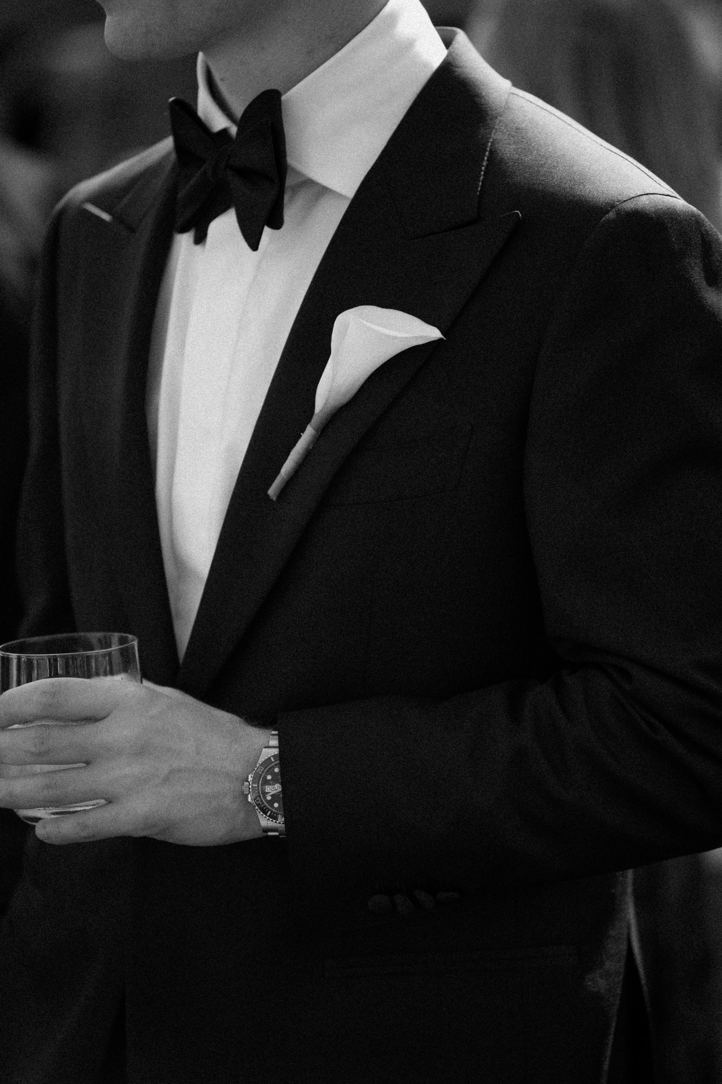 Man in tuxedo with bow tie and pocket square holding a glass, wearing a watch, at a formal event.