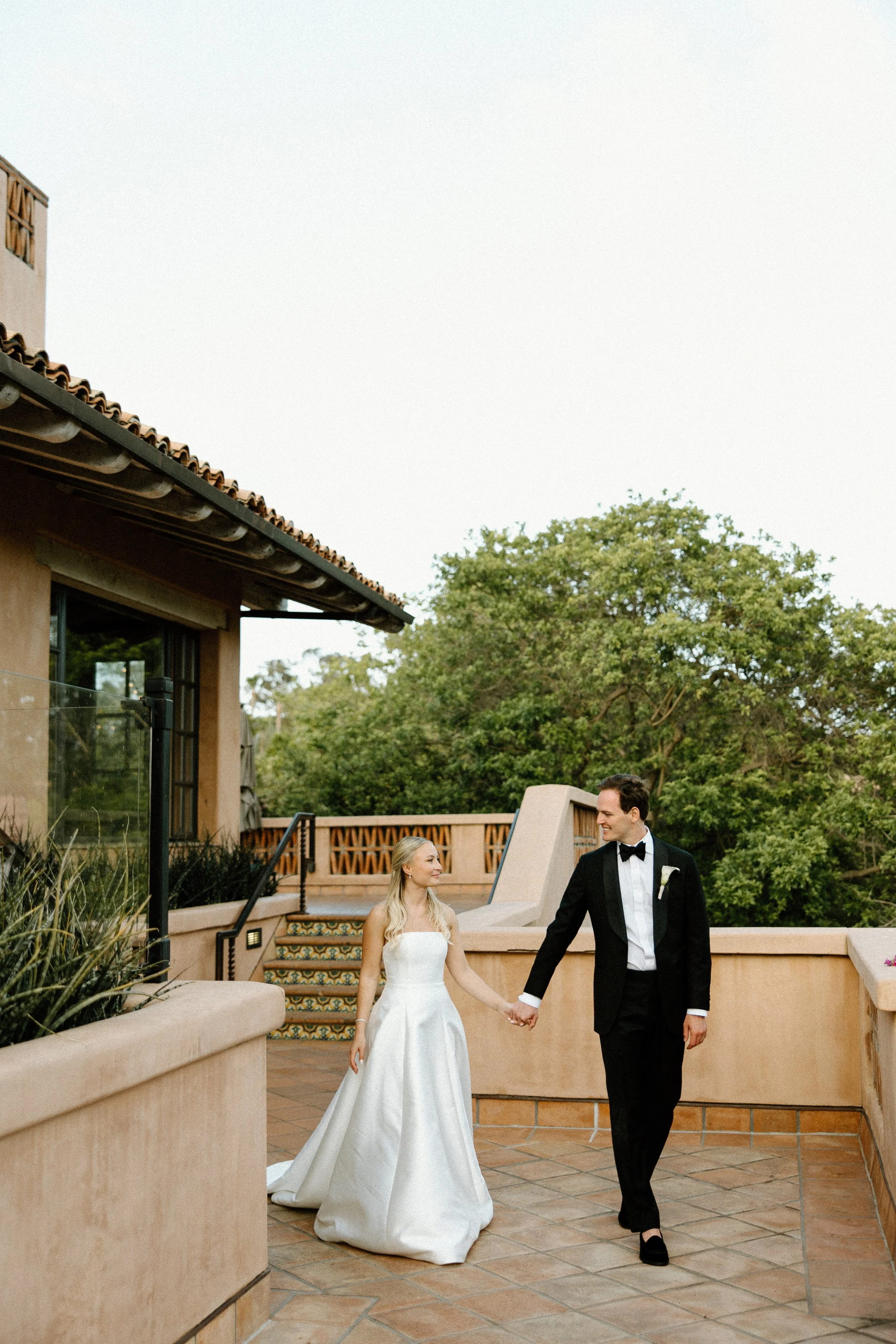 A bride in a white wedding gown and a groom in a black tuxedo walk hand in hand on a terrace with trees in the background, celebrating their wedding.