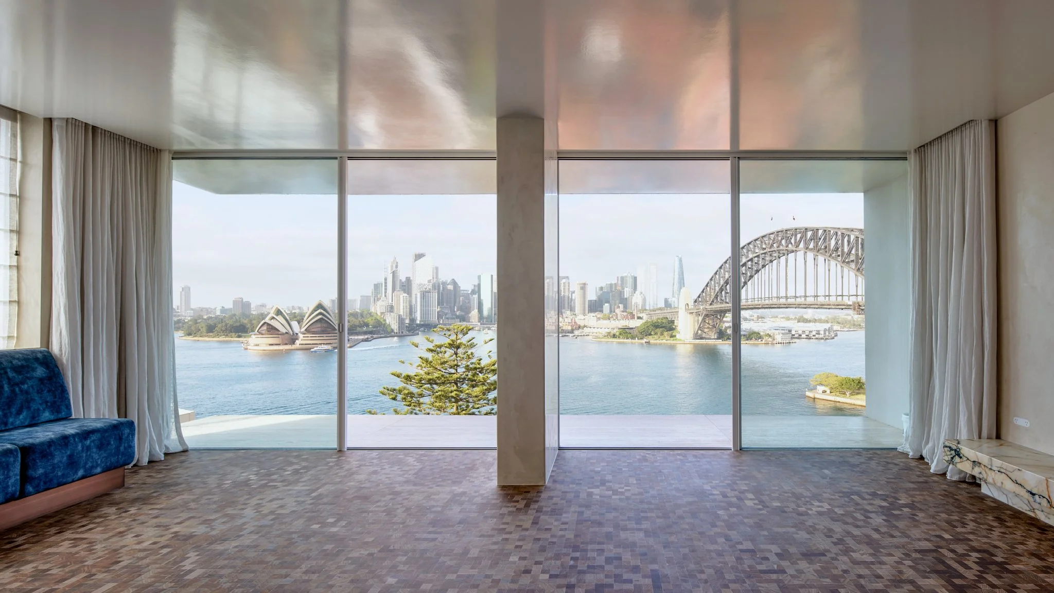 Room with large floor-to-ceiling windows overlooking Sydney Harbour, featuring the Sydney Opera House and Harbour Bridge, with a blue sofa on a wooden floor.