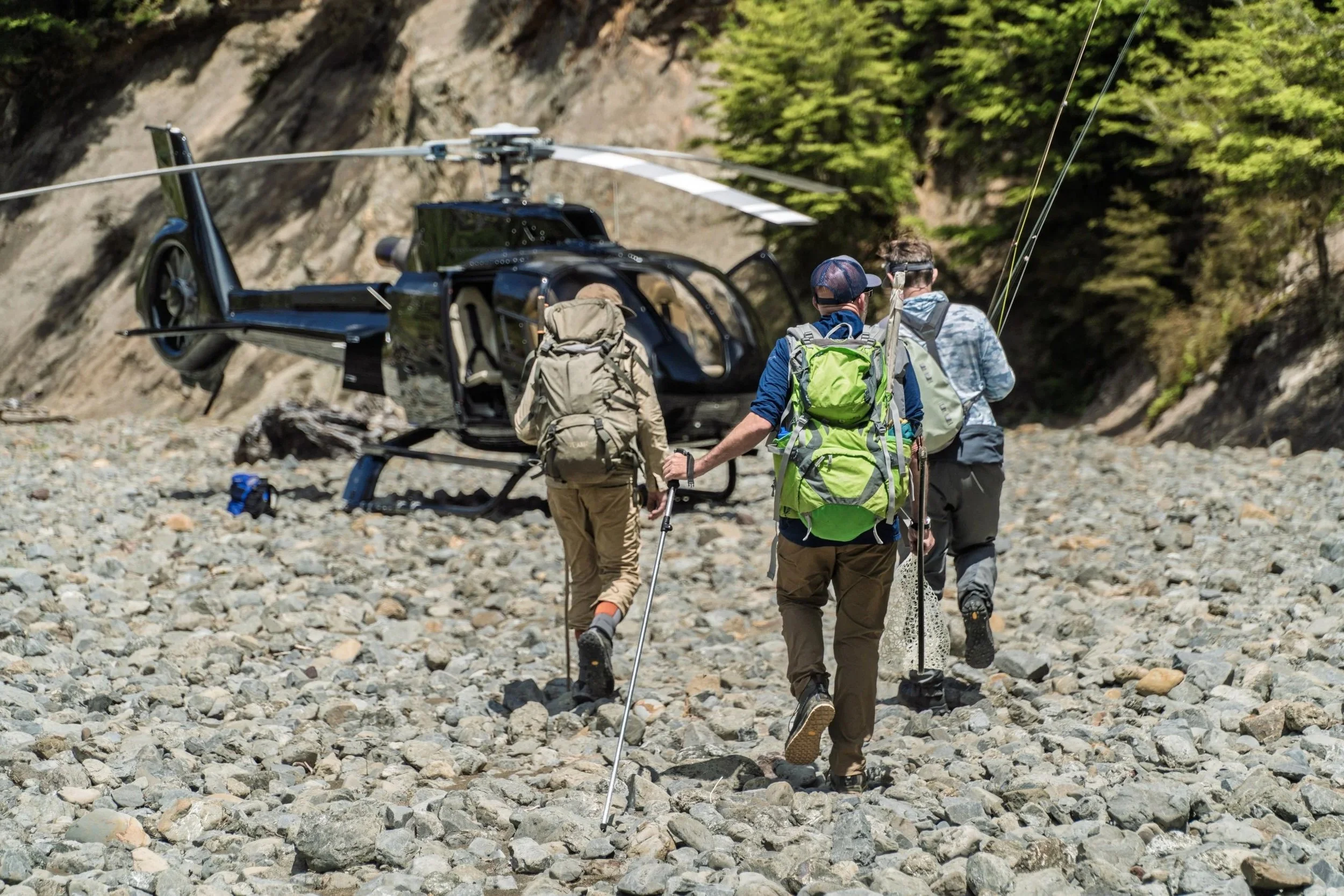 Four people with backpacks walk across a rocky riverbed, approaching a black helicopter with sliding doors open, surrounded by trees and mountain terrain.