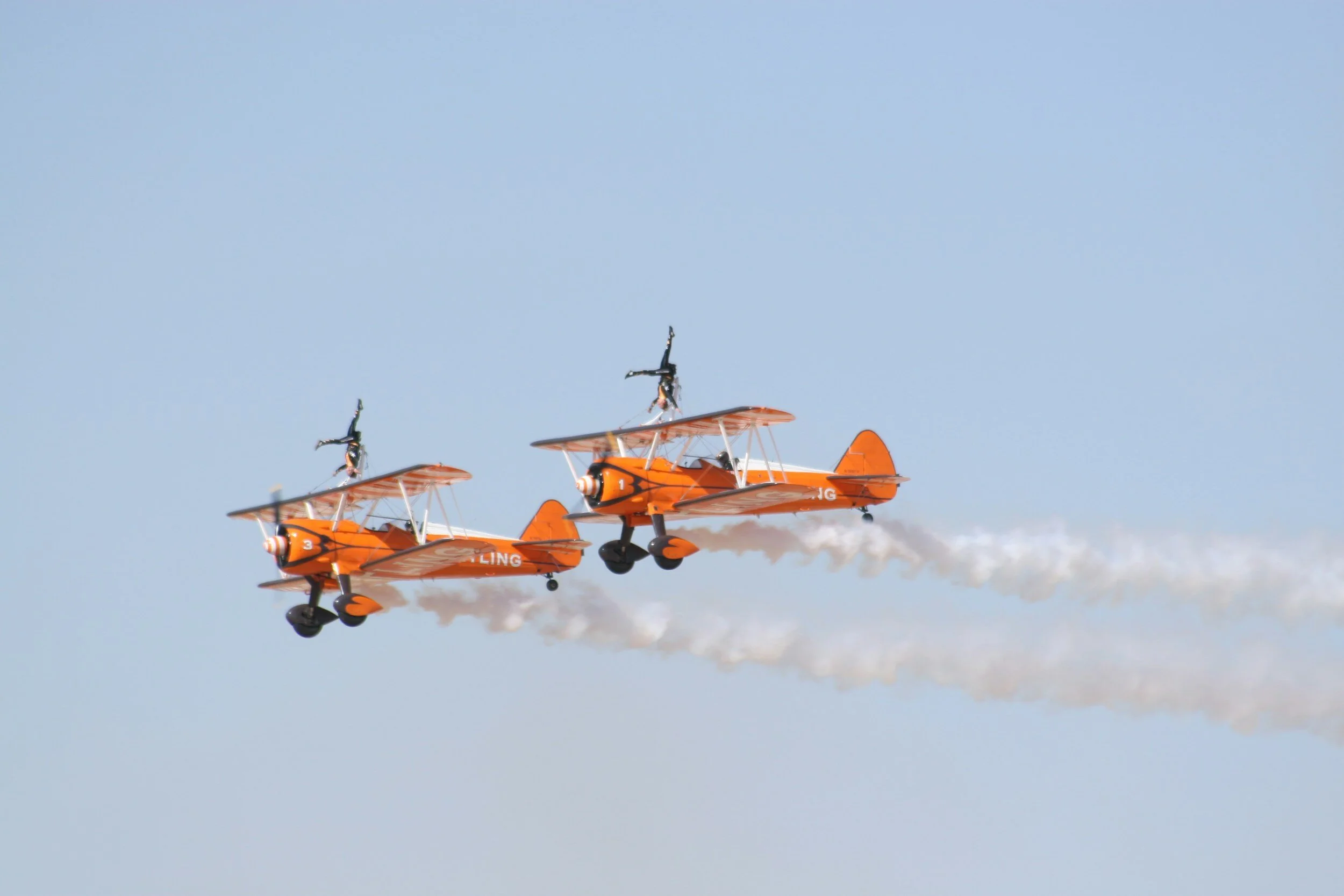 Two orange biplanes flying in formation with wing walkers performing stunts, leaving smoke trails against a clear blue sky.