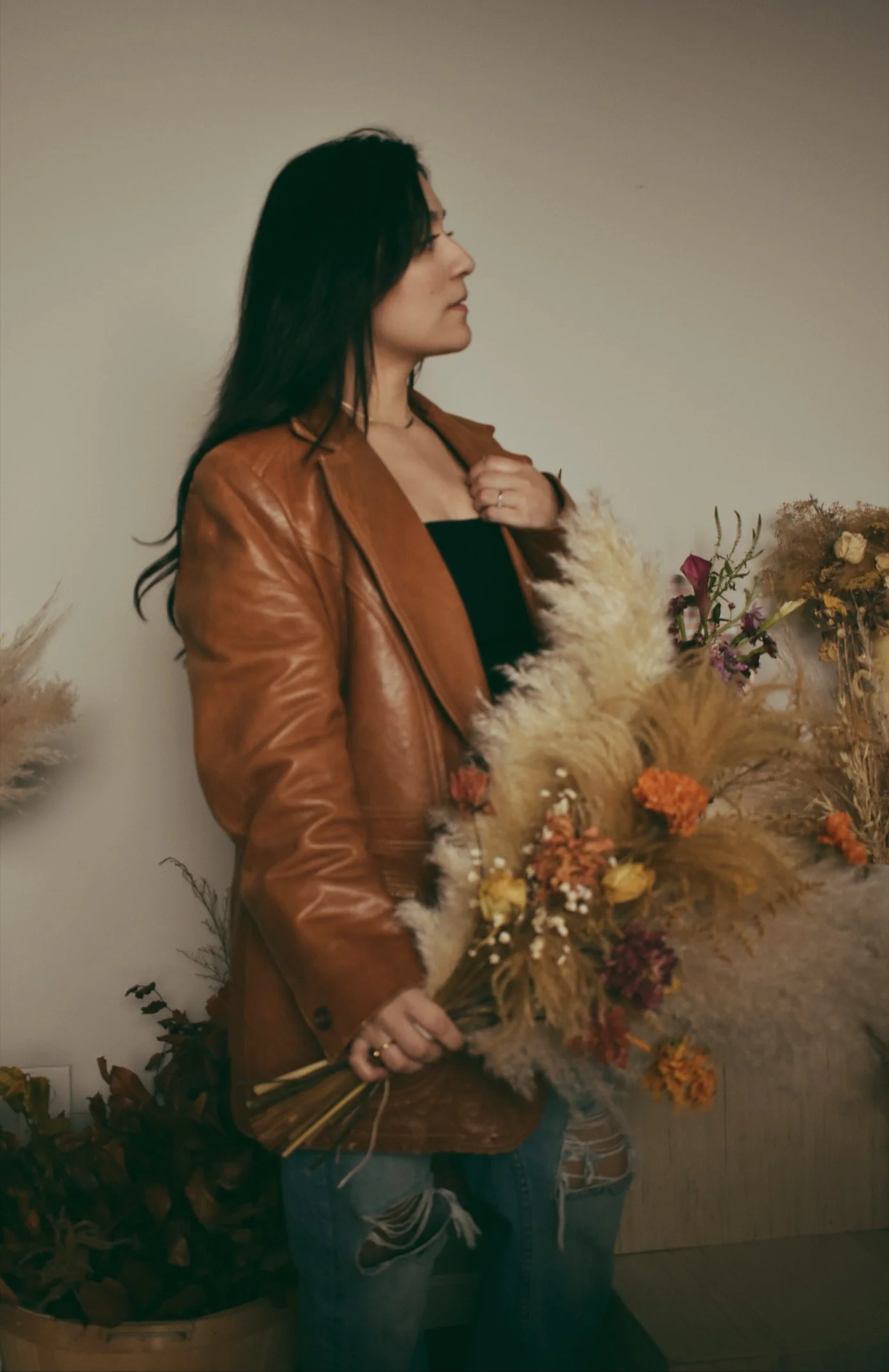 A woman with long dark hair wearing a brown leather jacket and ripped jeans, holding a bouquet of dried flowers, standing against a plain beige wall.