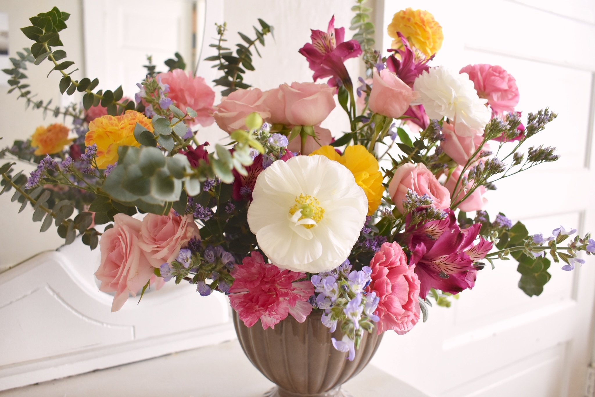 A large bouquet of colorful mixed flowers in a brown glass vase on a white surface with a white wall background.