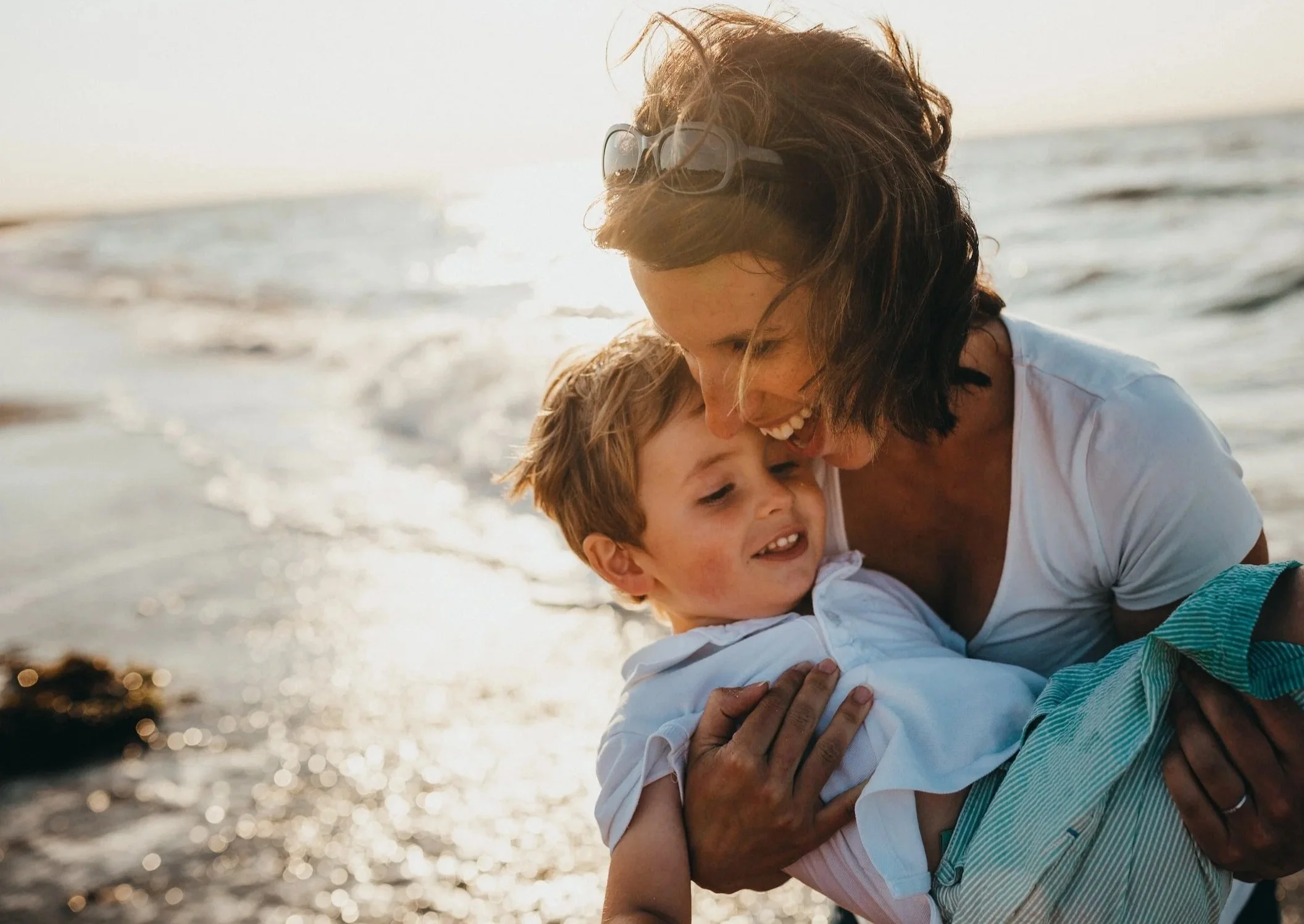 A woman and a young boy playing joyfully on a beach at sunset, with the woman holding the boy in her arms as they smile and laugh.