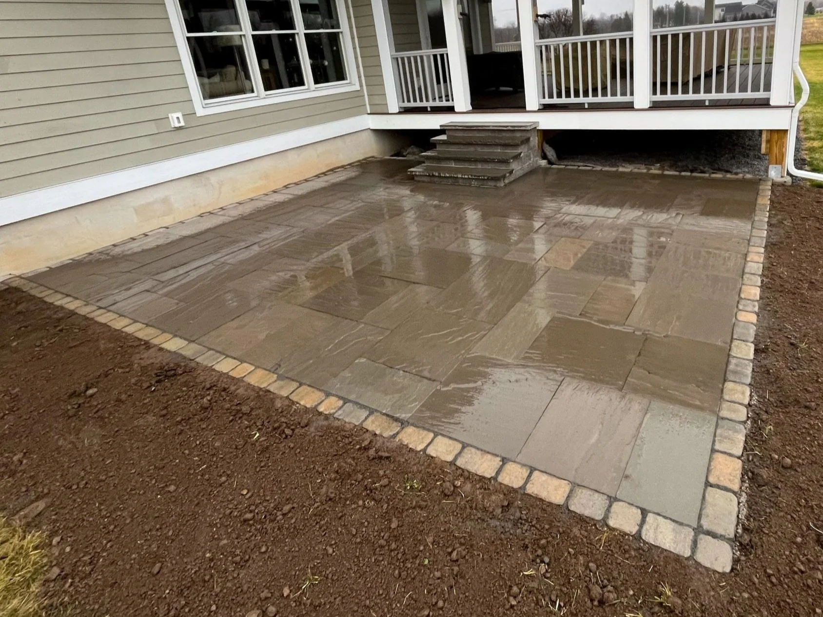Wet newly installed natural stone patio in front of a house with stairs leading to a deck, surrounded by dirt.