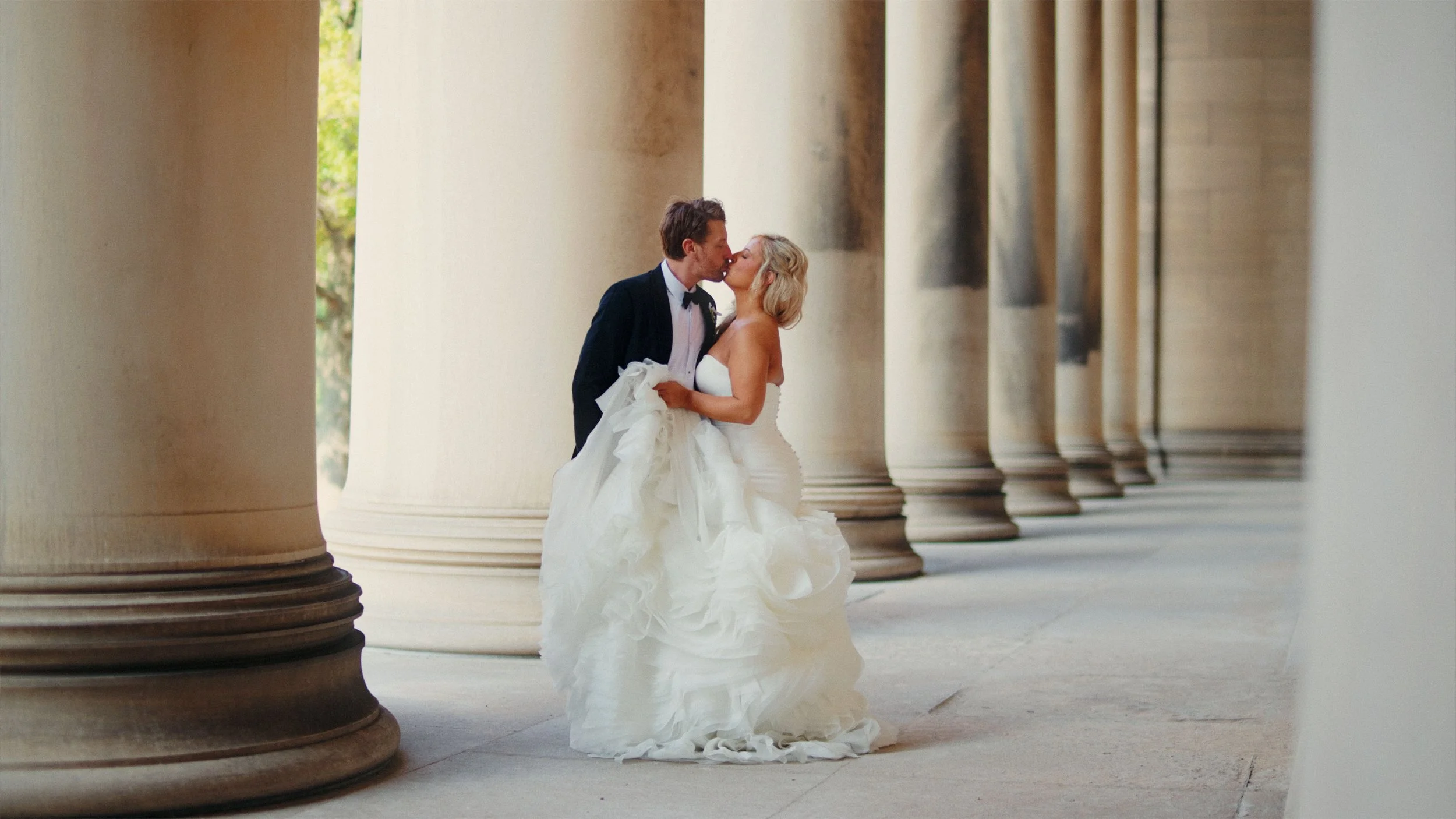 A bride and groom kiss among large stone columns, with the bride wearing a strapless white wedding gown and the groom in a tuxedo.