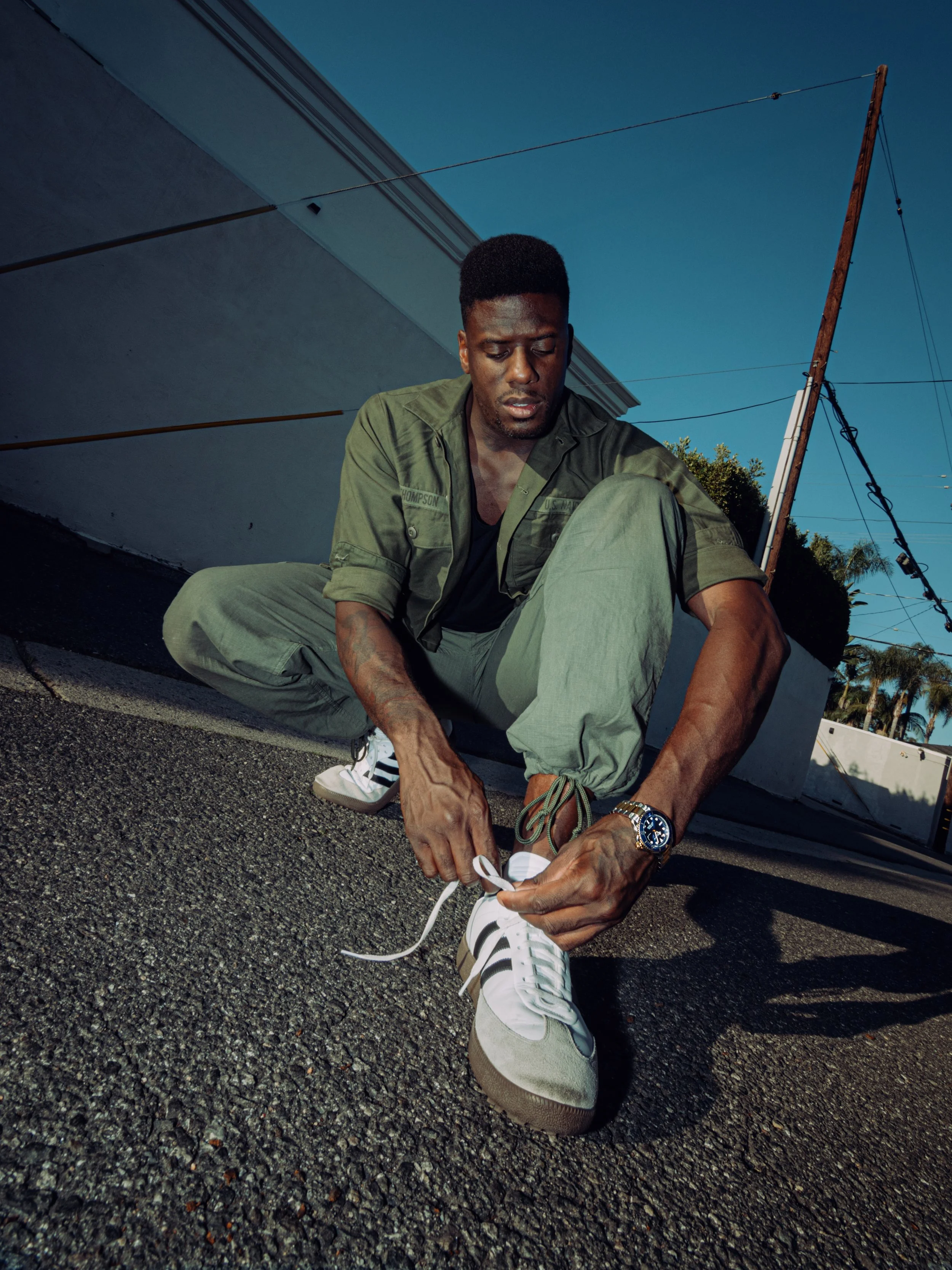 A man sitting on the ground outdoors, tying his shoe. He's wearing a green jacket, gray pants, and white sneakers with black stripes. The background features a pole with wires and a building, with a clear blue sky overhead.