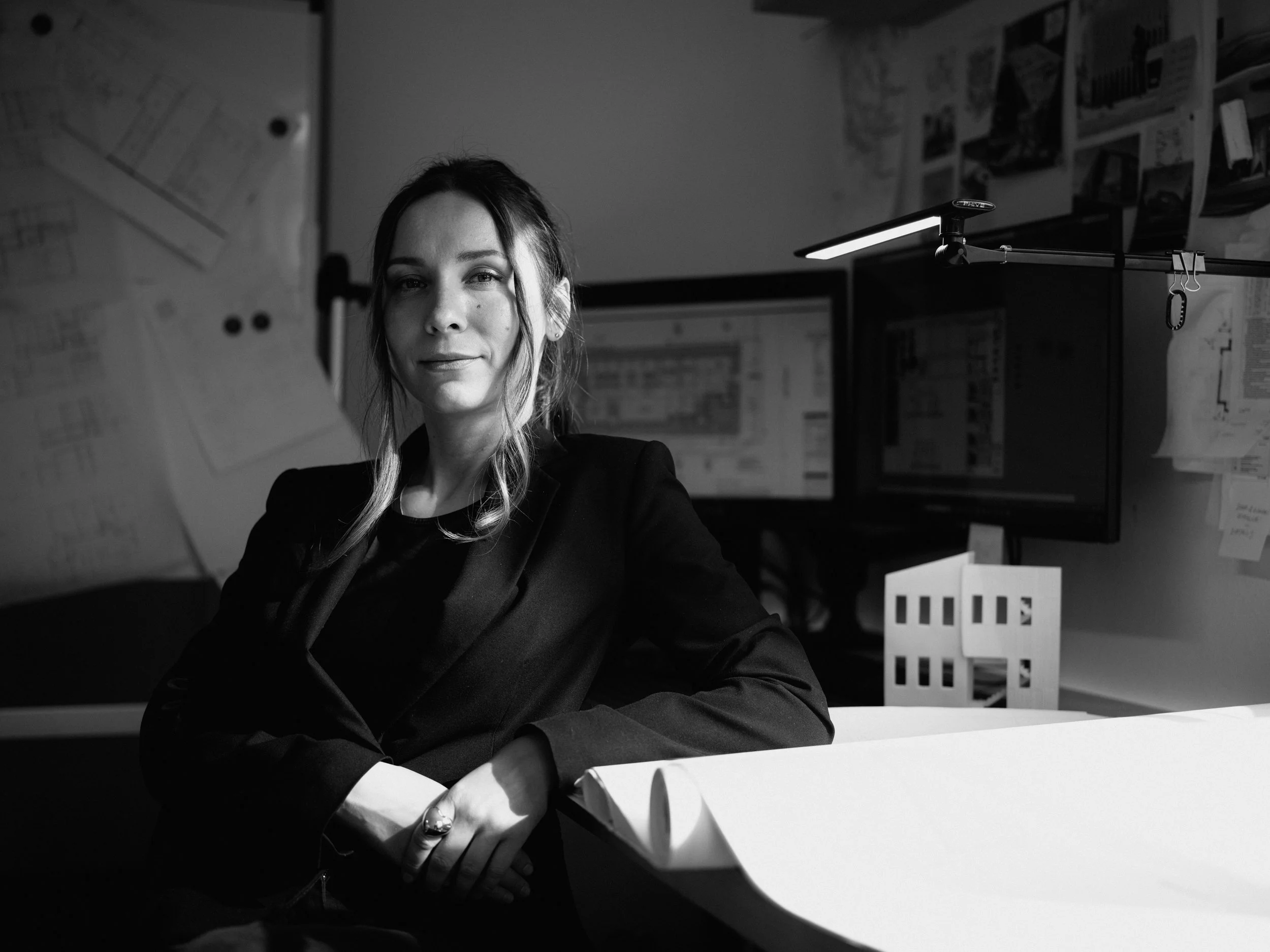 Black and white photo of a woman sitting at a desk in an office with multiple computer monitors and papers pinned to the wall behind her.