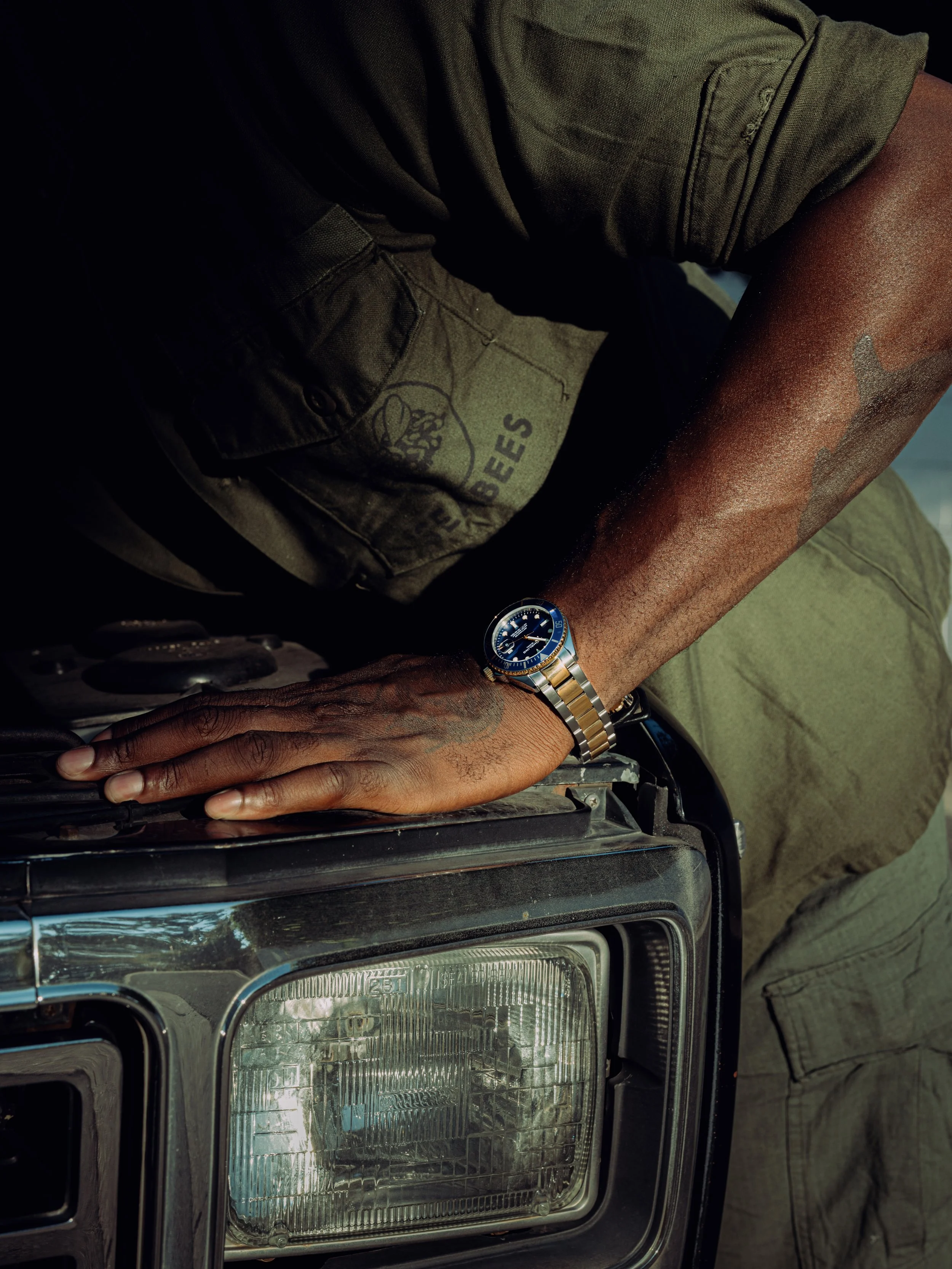 A person with a tattoo on their arm wearing a blue-faced wristwatch, leaning on the hood of a vintage black car.