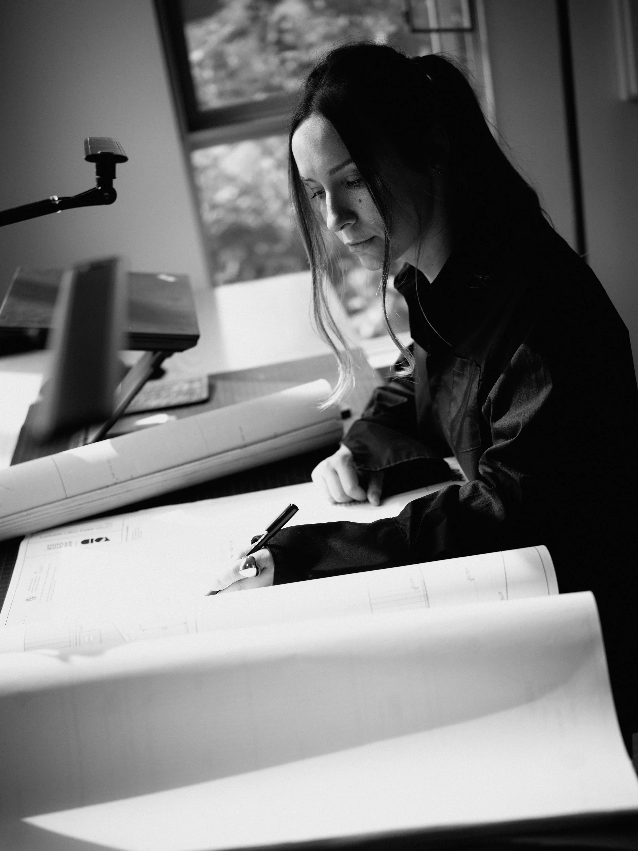 A woman in a dark jacket working with large rolled-up technical drawings or blueprints at a desk, in an office with a window showing trees outside.