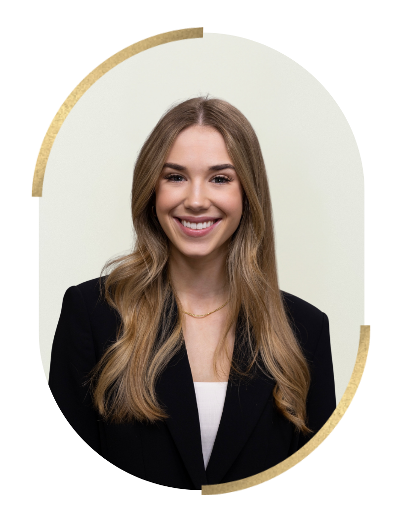 A young woman with long, wavy blonde hair wearing a black blazer, white top, and a gold necklace, smiling against a plain light-colored background.