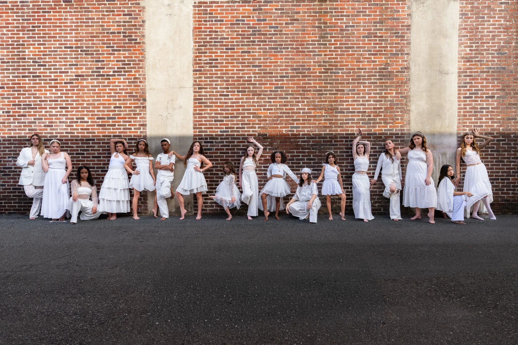 Group of young girls and women dressed in white poses against a brick wall outdoors.