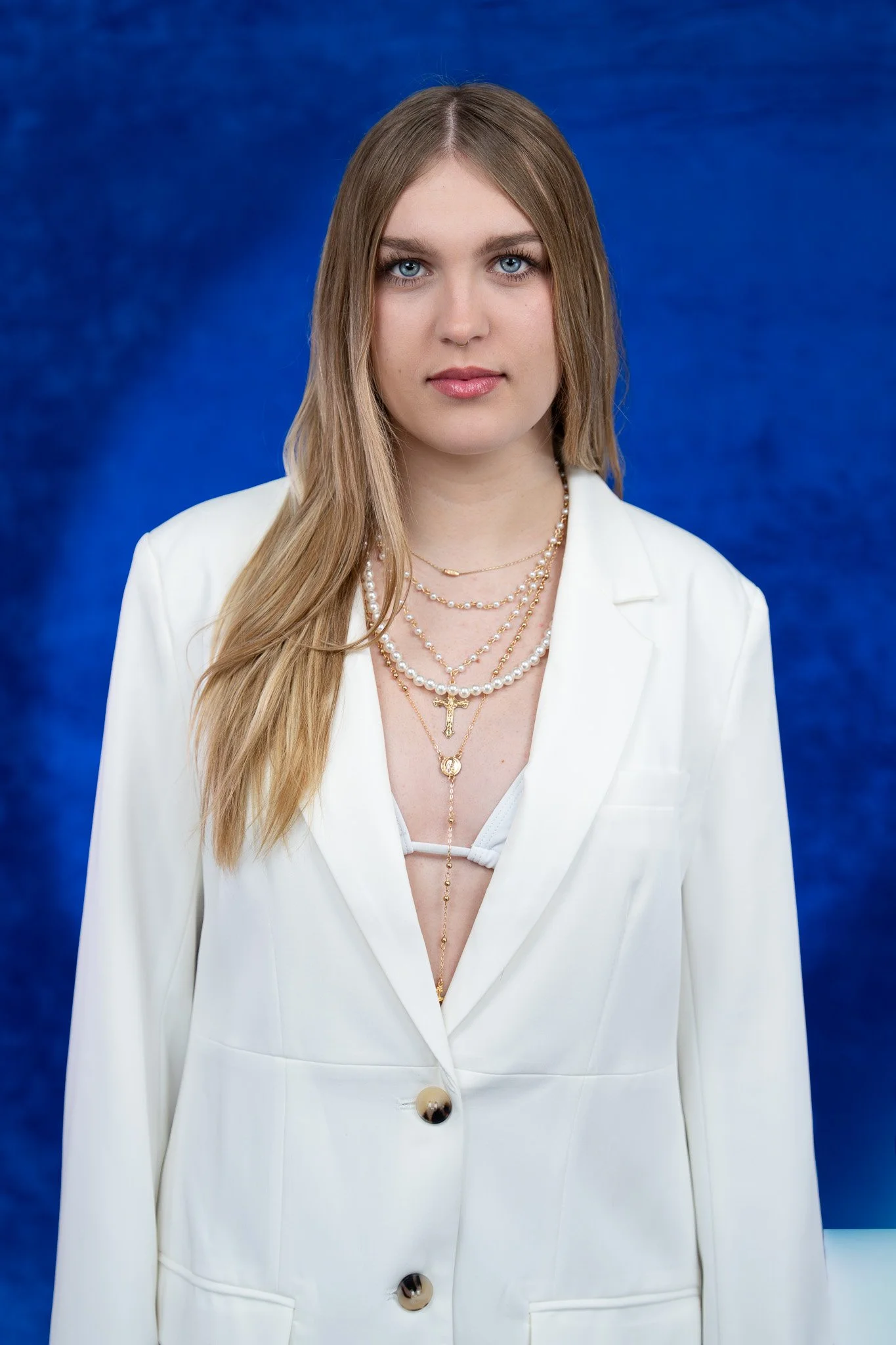 A woman with long blonde hair and blue eyes wearing a white blazer and layered necklaces, standing against a blue background.