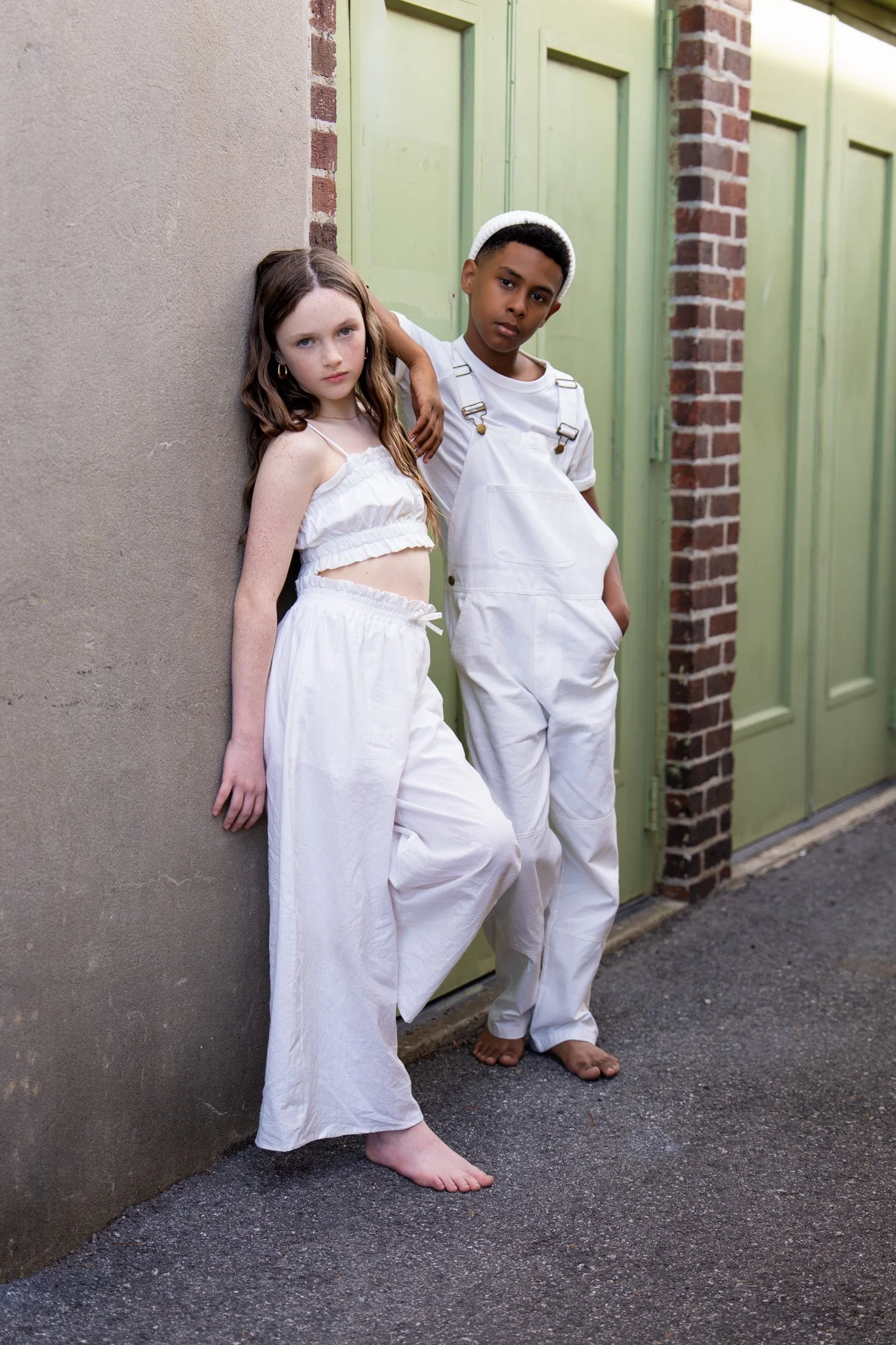 Two children, a girl and a boy, standing barefoot on this sidewalk against a beige wall and green door, with the girl leaning her back against the wall and the boy resting his arm on her shoulder.