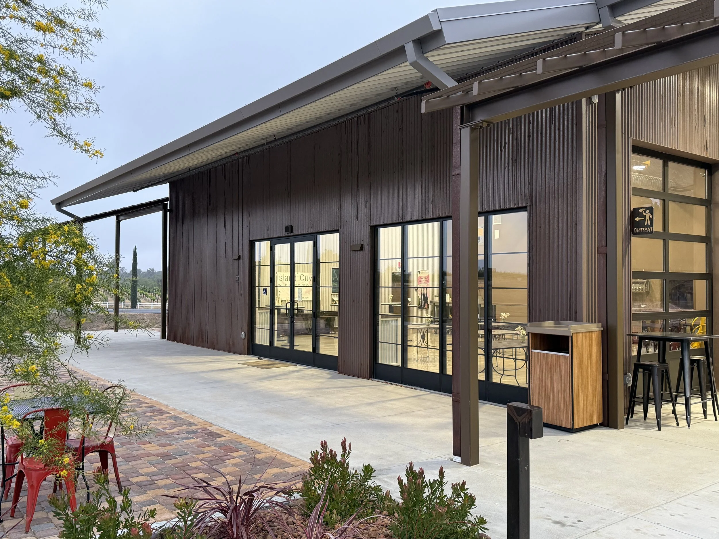 Exterior of a modern building with large glass doors and windows, wooden siding, outdoor seating area with high tables and chairs, and landscaped plants in the foreground.