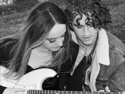 Two young women sitting close together, one playing an acoustic guitar, outdoors.