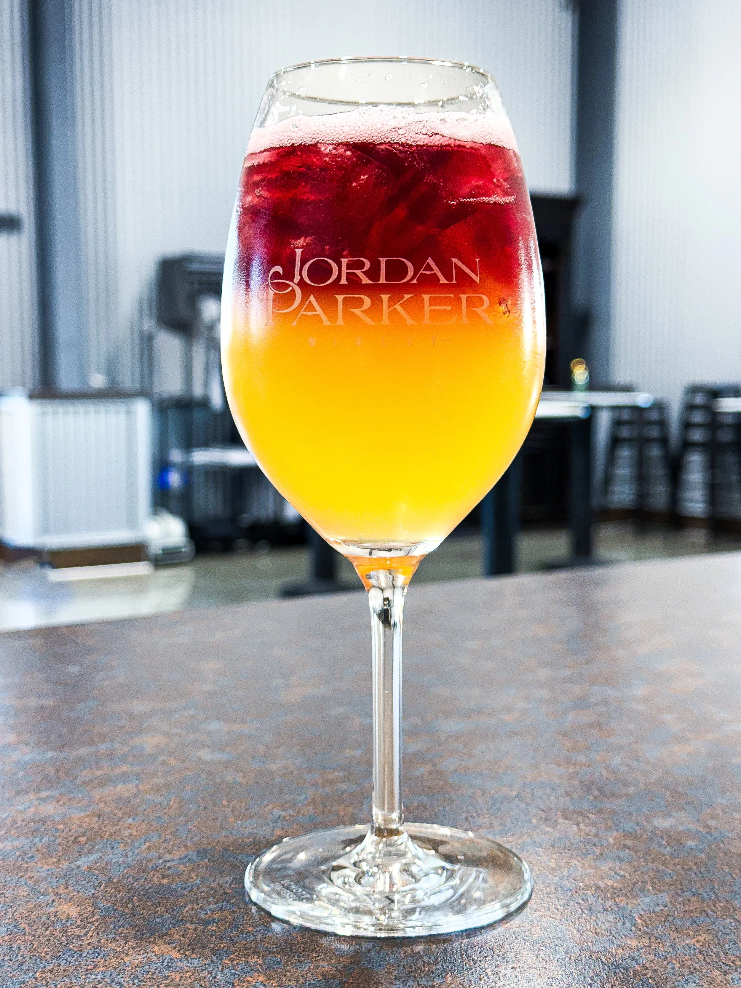 A glass of layered beverage with red on top and yellow at the bottom, with the name 'Jordan Parker' printed on the glass, placed on a speckled countertop.