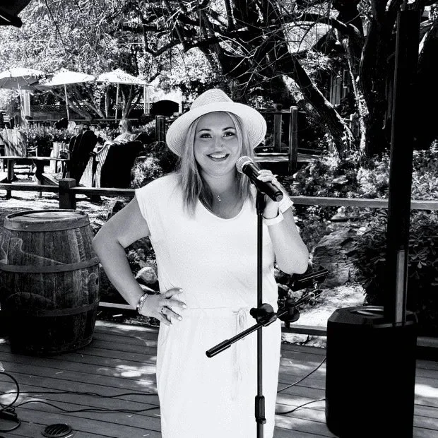 A woman wearing a white dress and a wide-brimmed hat, standing outdoors with a microphone, smiling. There are trees, outdoor seating, and barrels in the background.