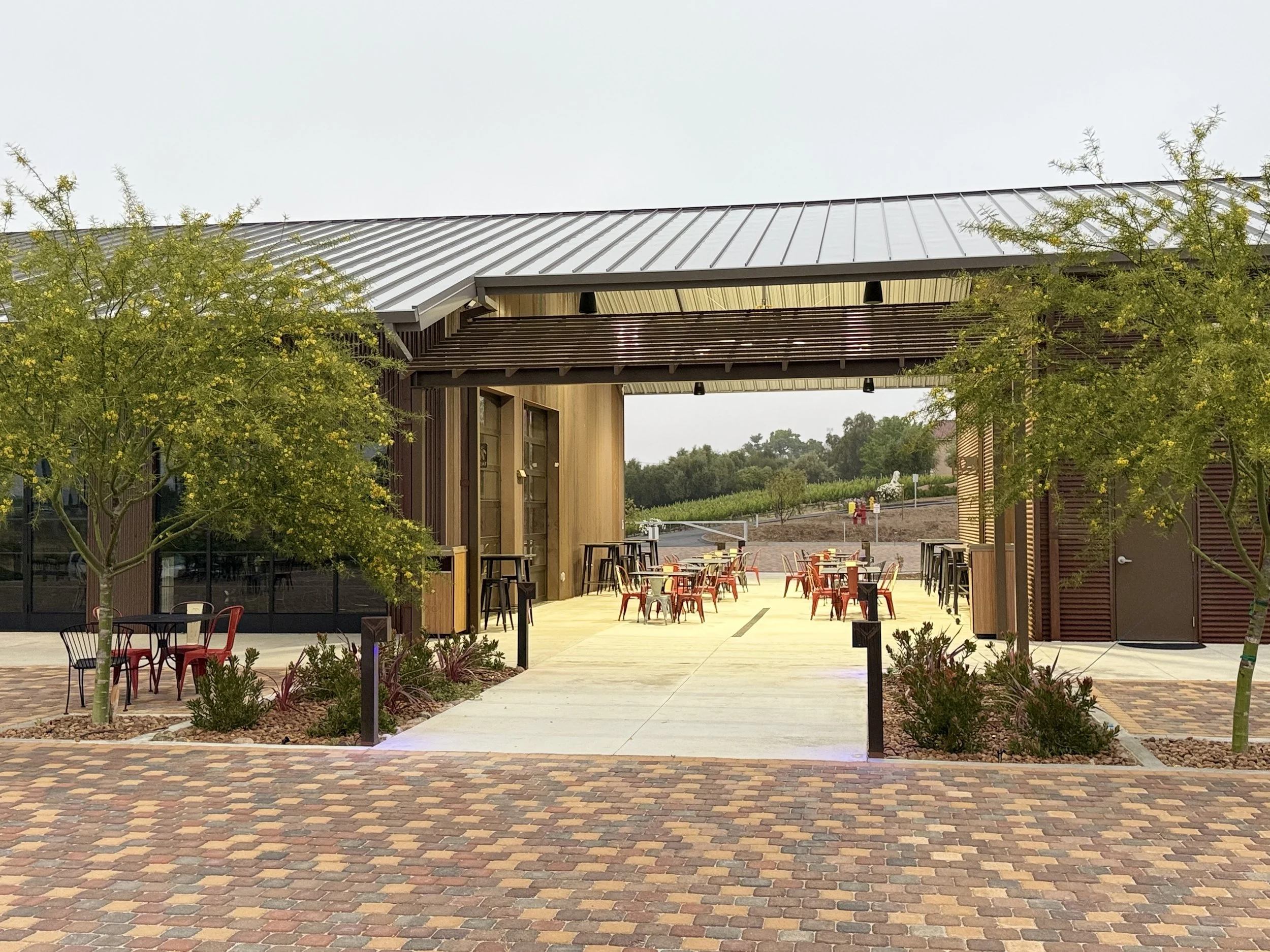 Modern outdoor dining area with wooden and metal furniture, trees, and a brick pathway leading to a covered patio in a vineyard setting.