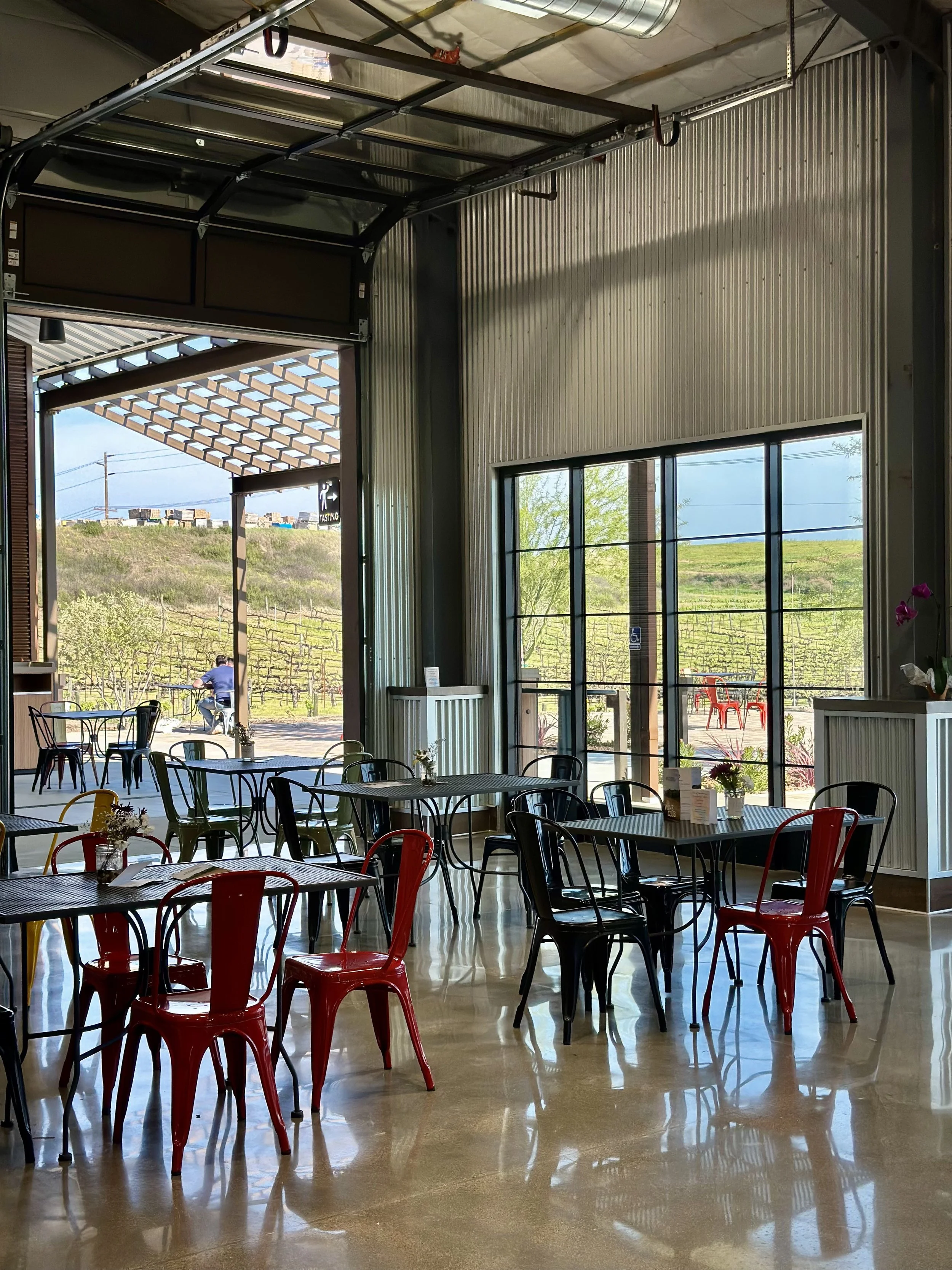 Interior of a cafe or restaurant with colorful chairs, tables with small flower arrangements, large windows, and a view of a vineyard outside.