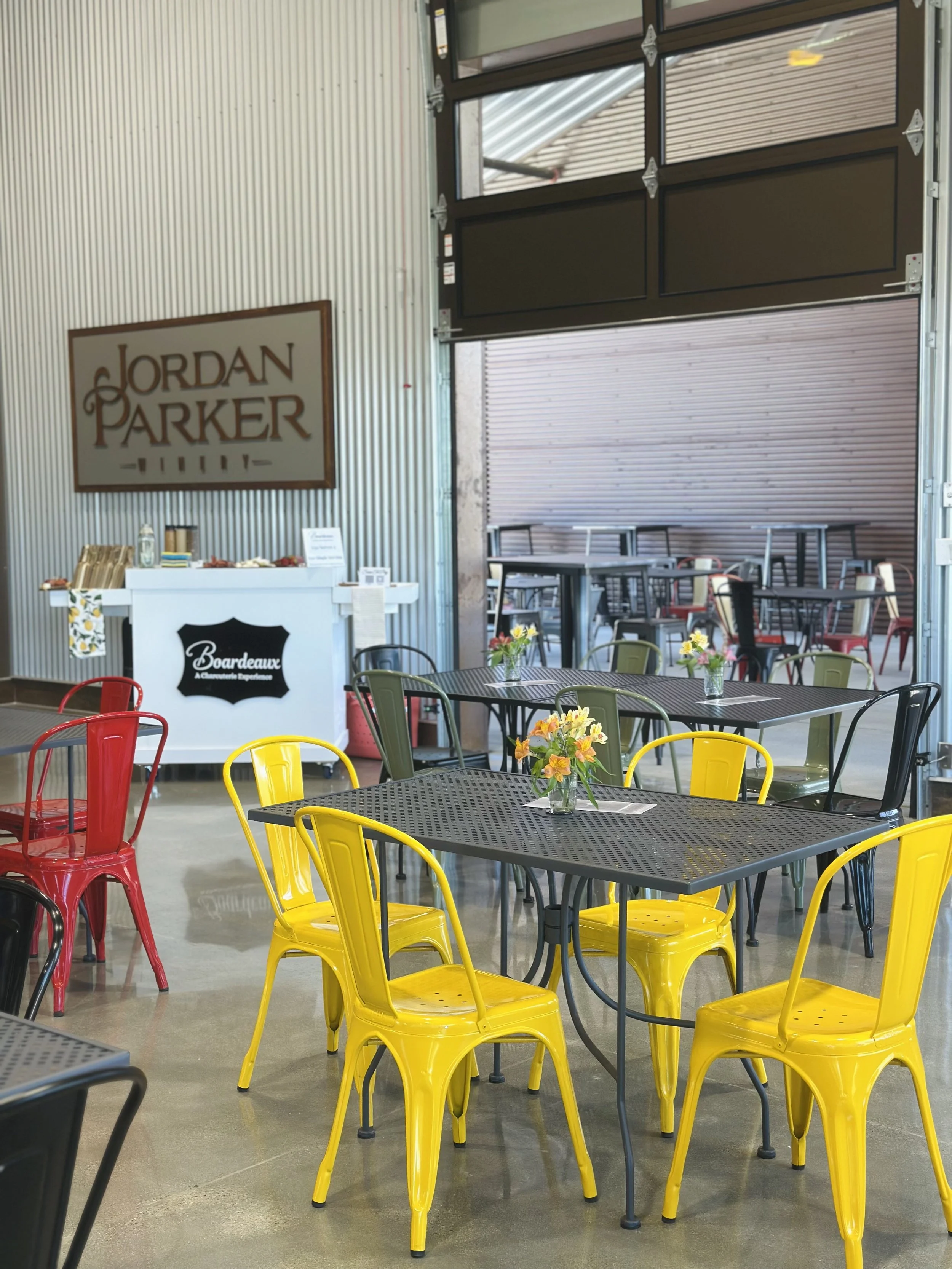 Indoor seating area with black, yellow, gray, and red metal chairs, tables with small flower arrangements, a dessert stand, and a partially open garage door in the background showing additional seating with tables and chairs.