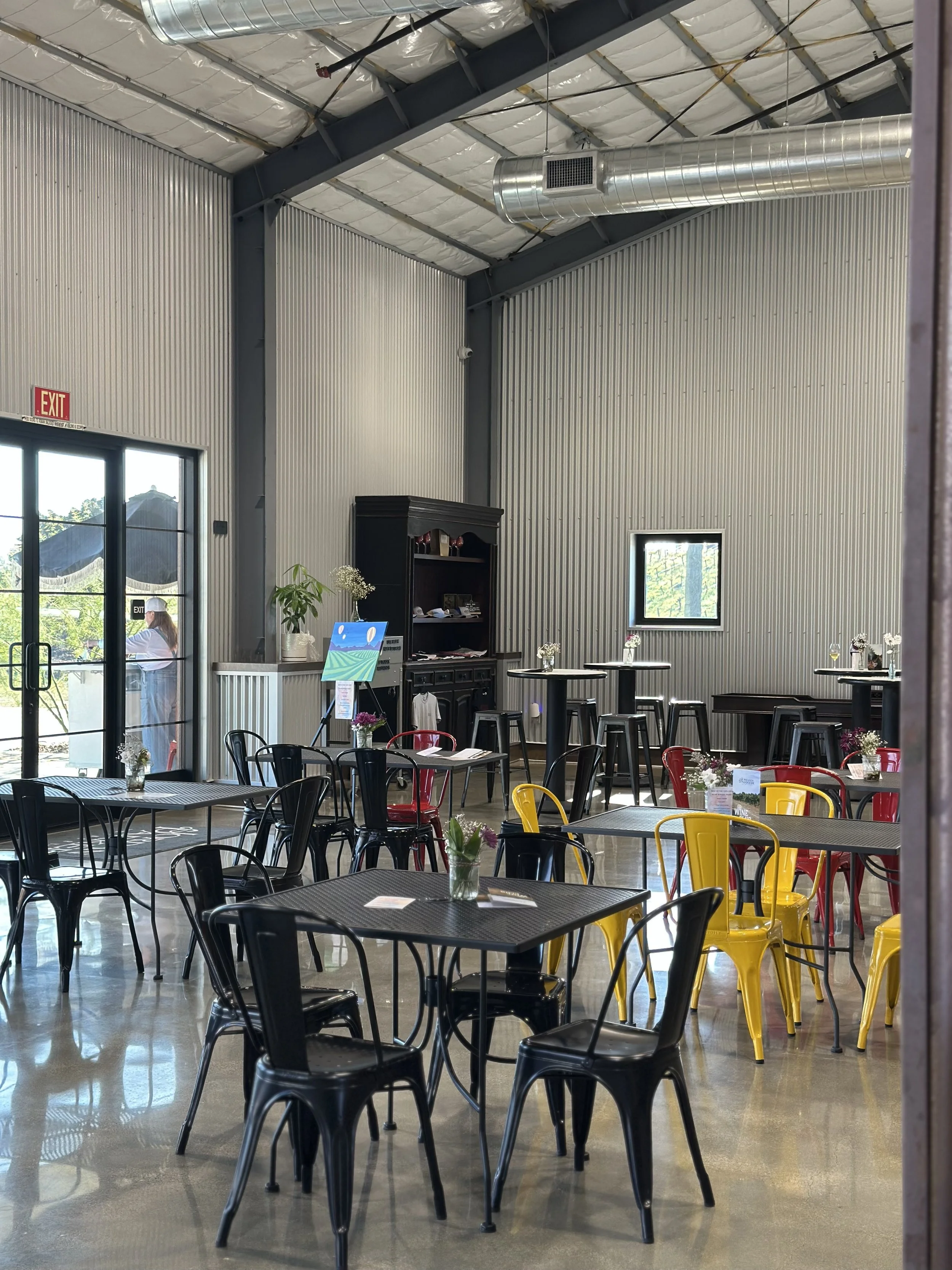 Interior of a modern cafe or restaurant with metal walls and ceiling, black, yellow, and red chairs, small tables with vases of flowers, and a serving area with black shelving. Large windows and glass door showing outside scenery.