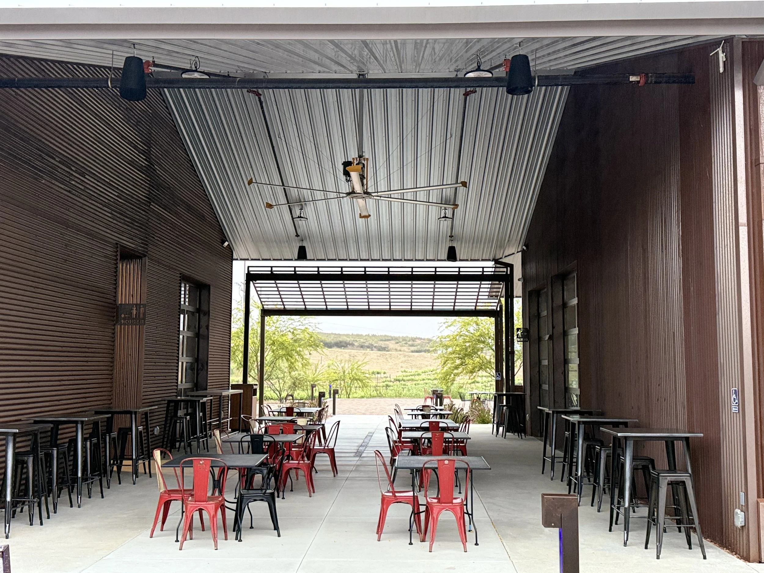 Empty outdoor dining area with black and red chairs and tables under a metal roof, open view of trees and fields in the background.