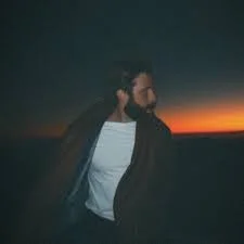 A man with a beard wearing a dark jacket and white T-shirt looking to the side at sunset over a horizon.