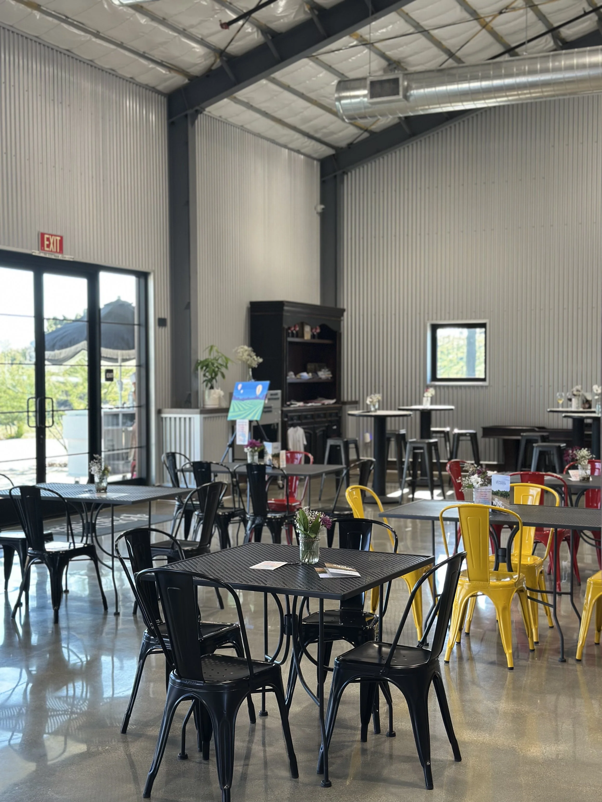 Interior of a modern cafe or restaurant with black and yellow chairs, black tables, and minimalistic decor. Sunlight coming through glass doors and windows, with plants and flowers on the tables.