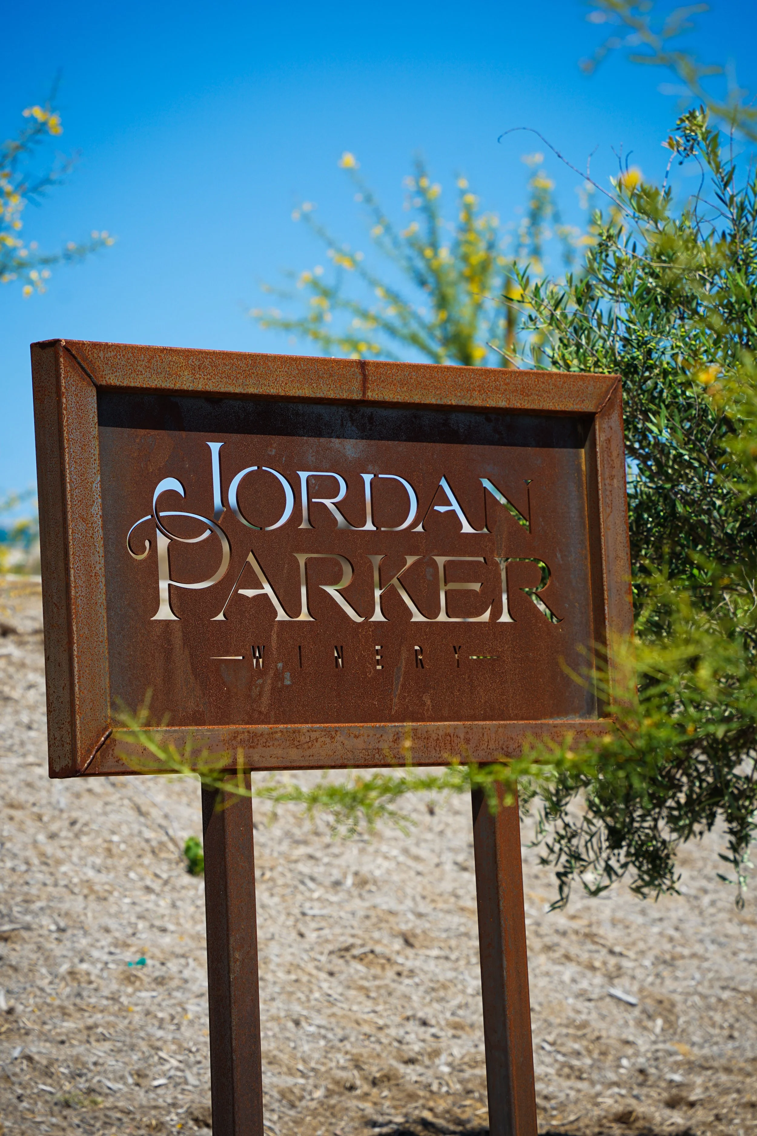 Rusty metal sign reading 'Jordan Parker Winery' with blue sky and green bushes in the background.