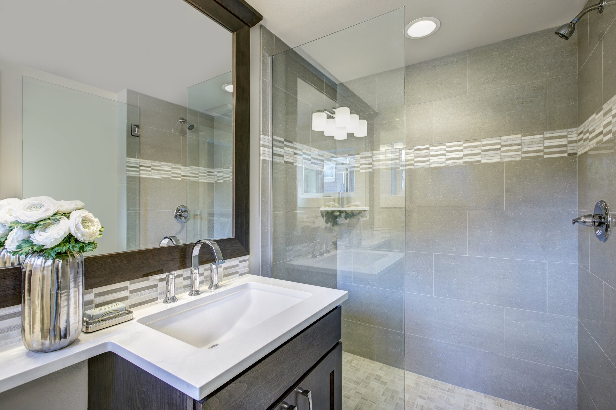Modern bathroom with tiled walk-in shower, white sink with chrome faucet, large mirror, and decorative vase with white flowers.