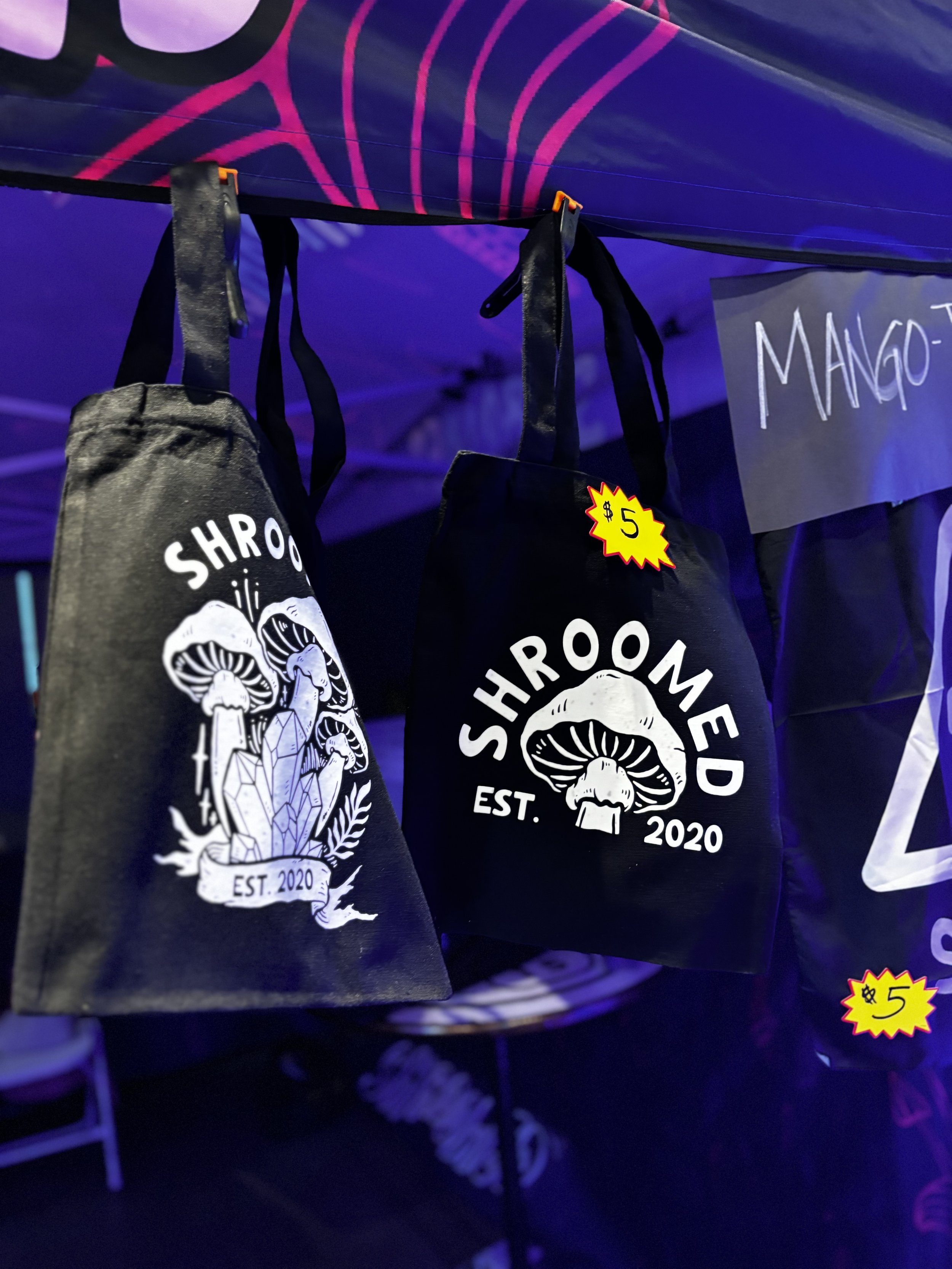 Black tote bags with a white mushroom and crystal design, the word 'SHROOMED', and 'EST. 2020' printed on them, hanging at a store stall with a sign showing $5.